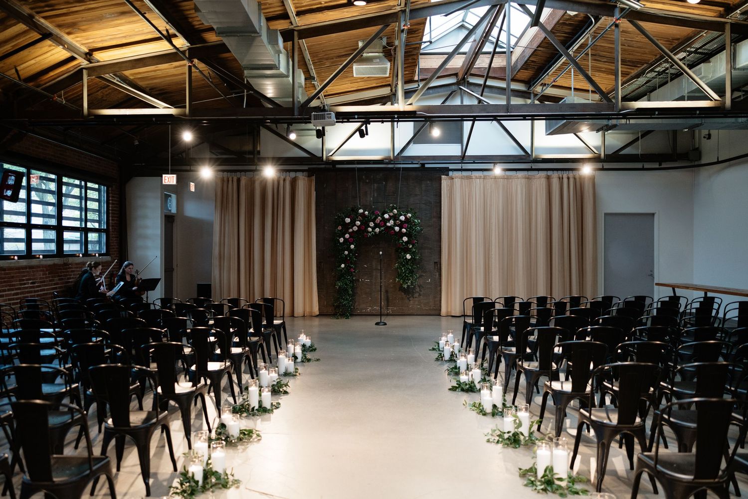 Modern industrial wedding ceremony space with rows of black chairs and exposed ceiling beams.