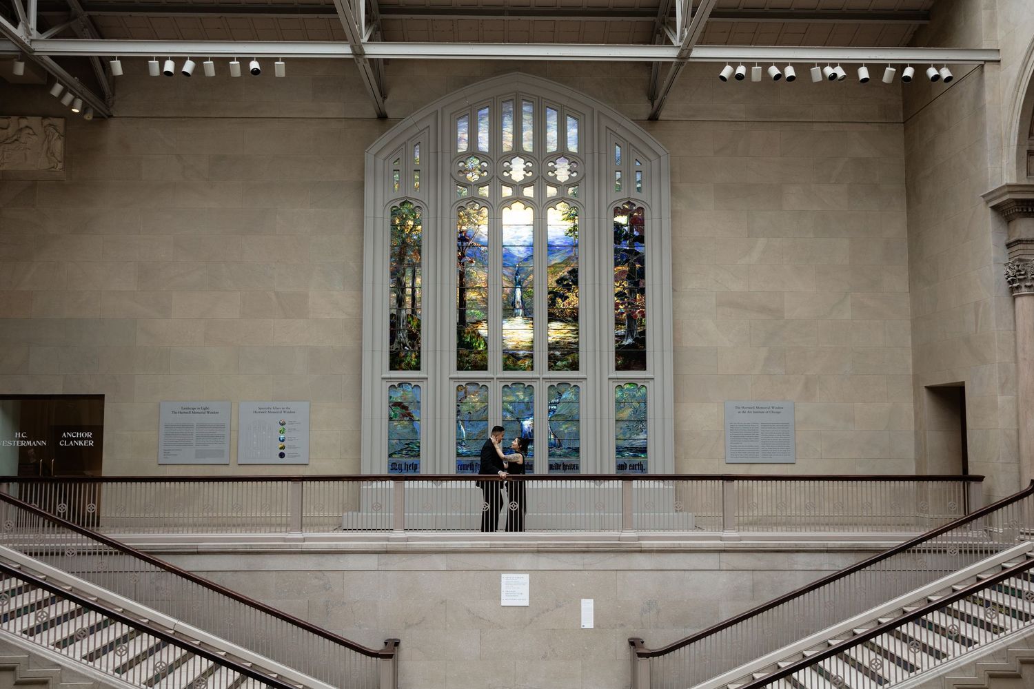 A couple stands before a large stained glass window in a grand historic building interior.