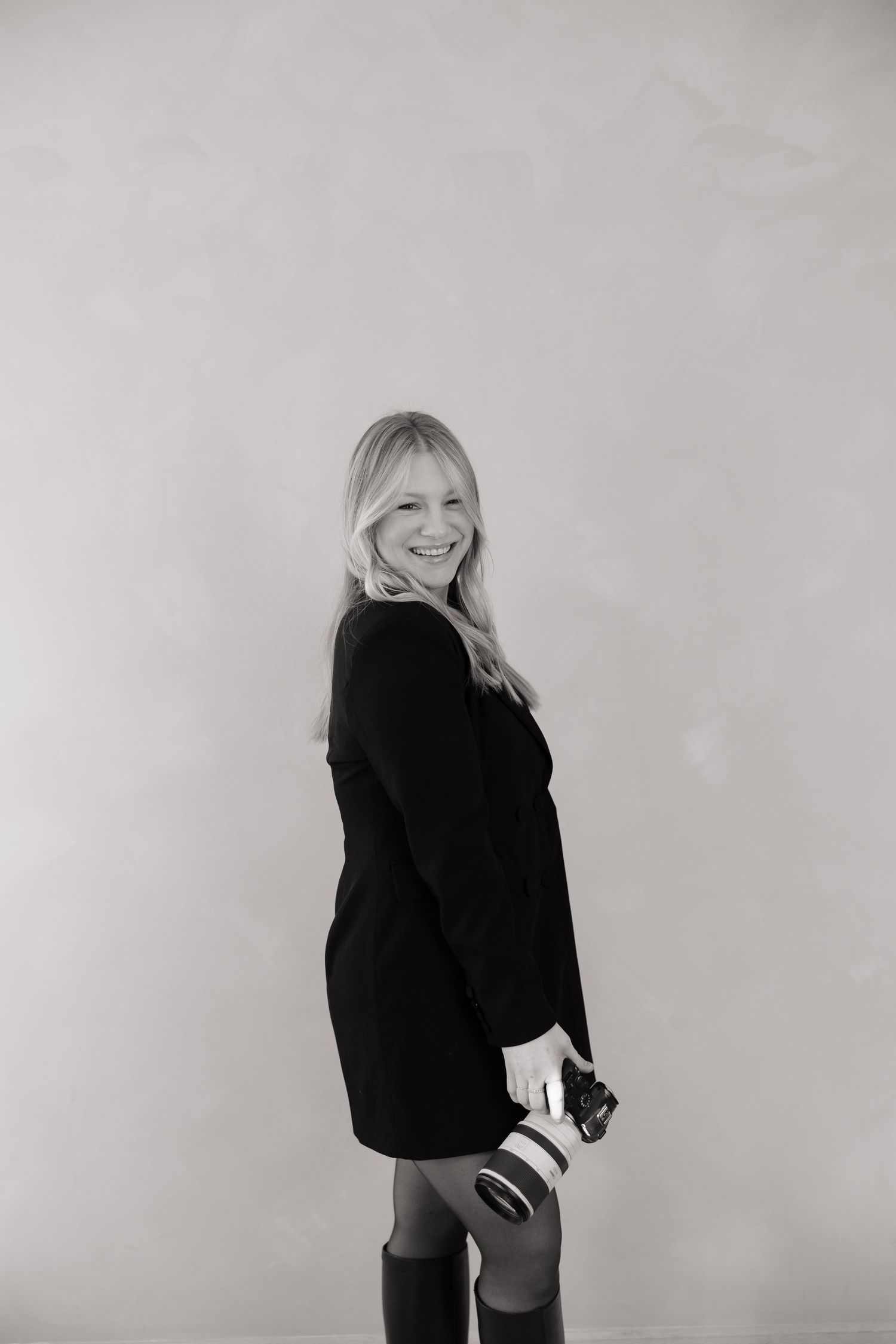 Black and white portrait of a person in a dark sweater dress and boots posing against a light background.