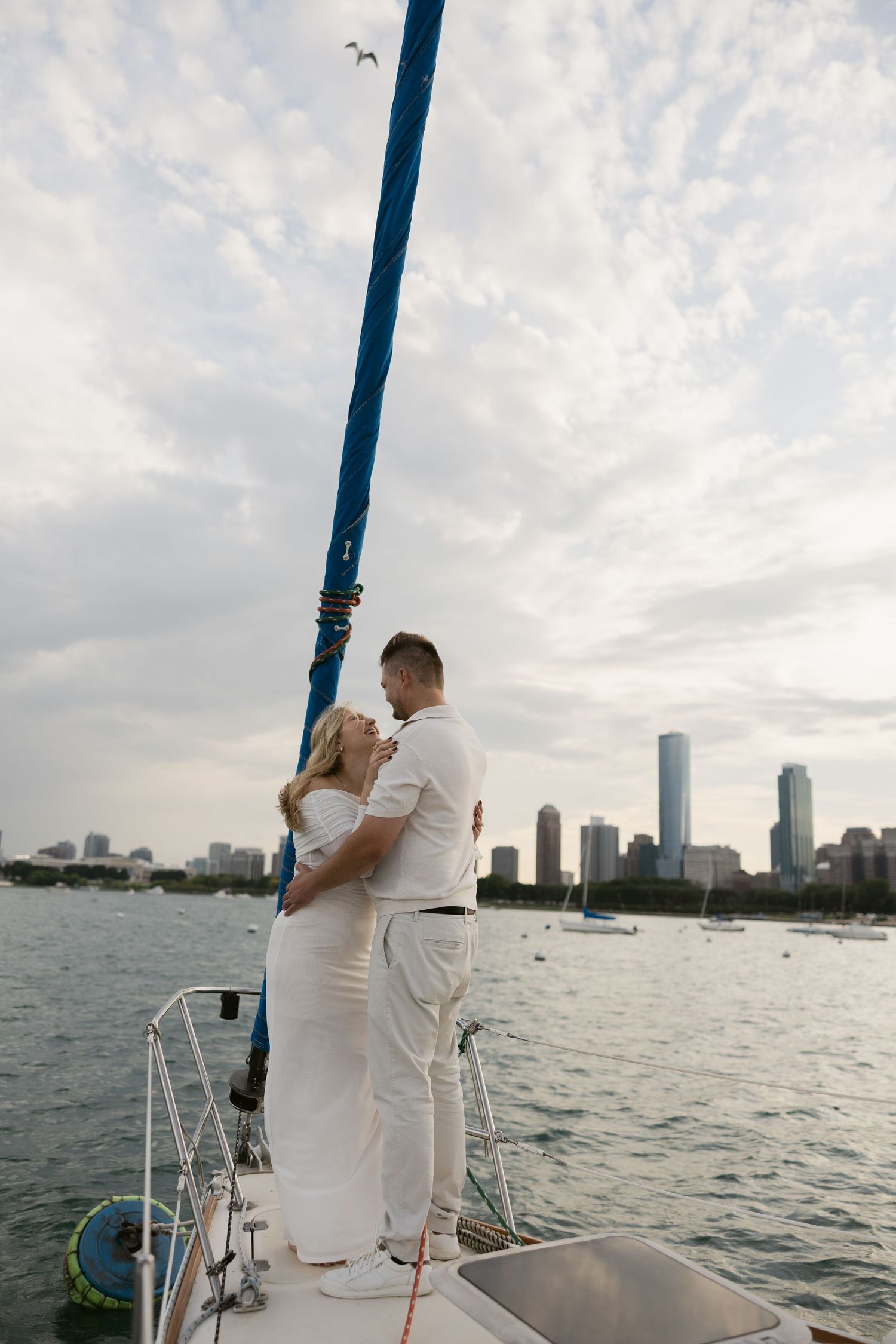 A couple in white outfits shares a romantic moment on a boat with a cityscape visible in the background.