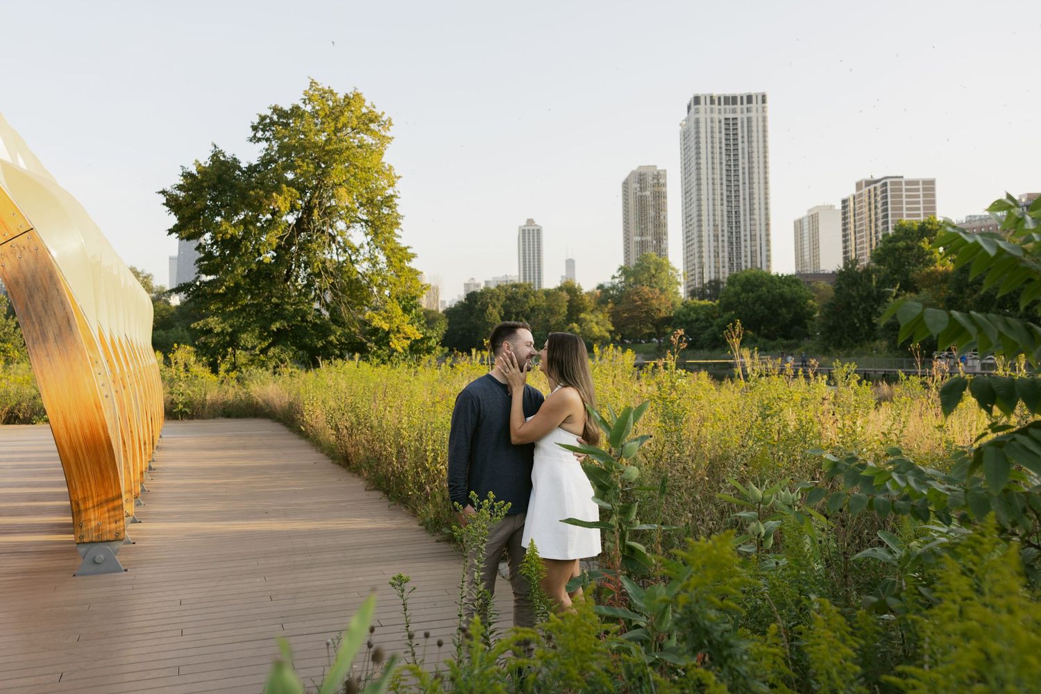 A couple enjoys a romantic sunset walk along a wooden boardwalk path through tall grass and wildflowers.