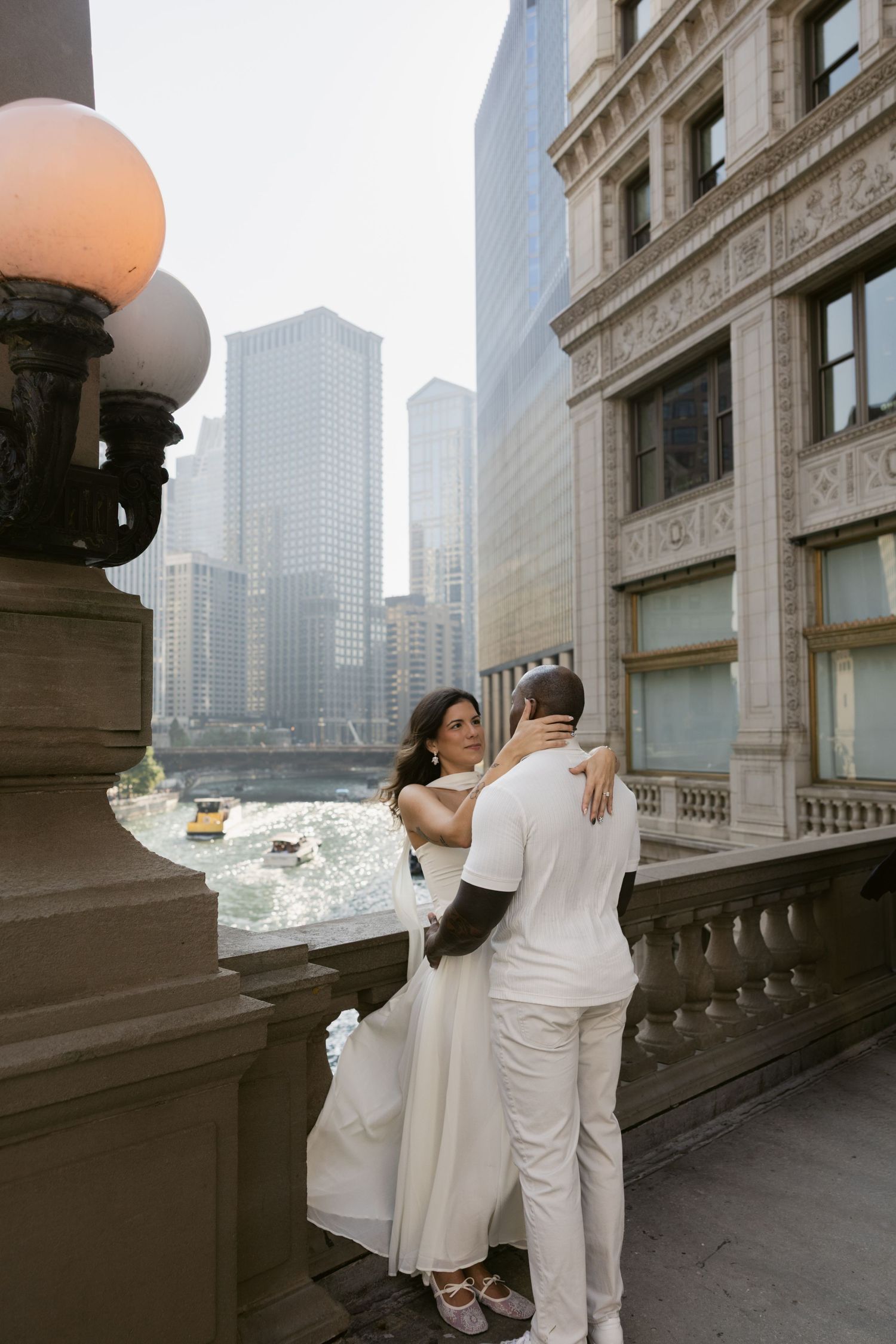 A couple in white outfits embraces on a stone balcony overlooking Chicago's urban skyline.