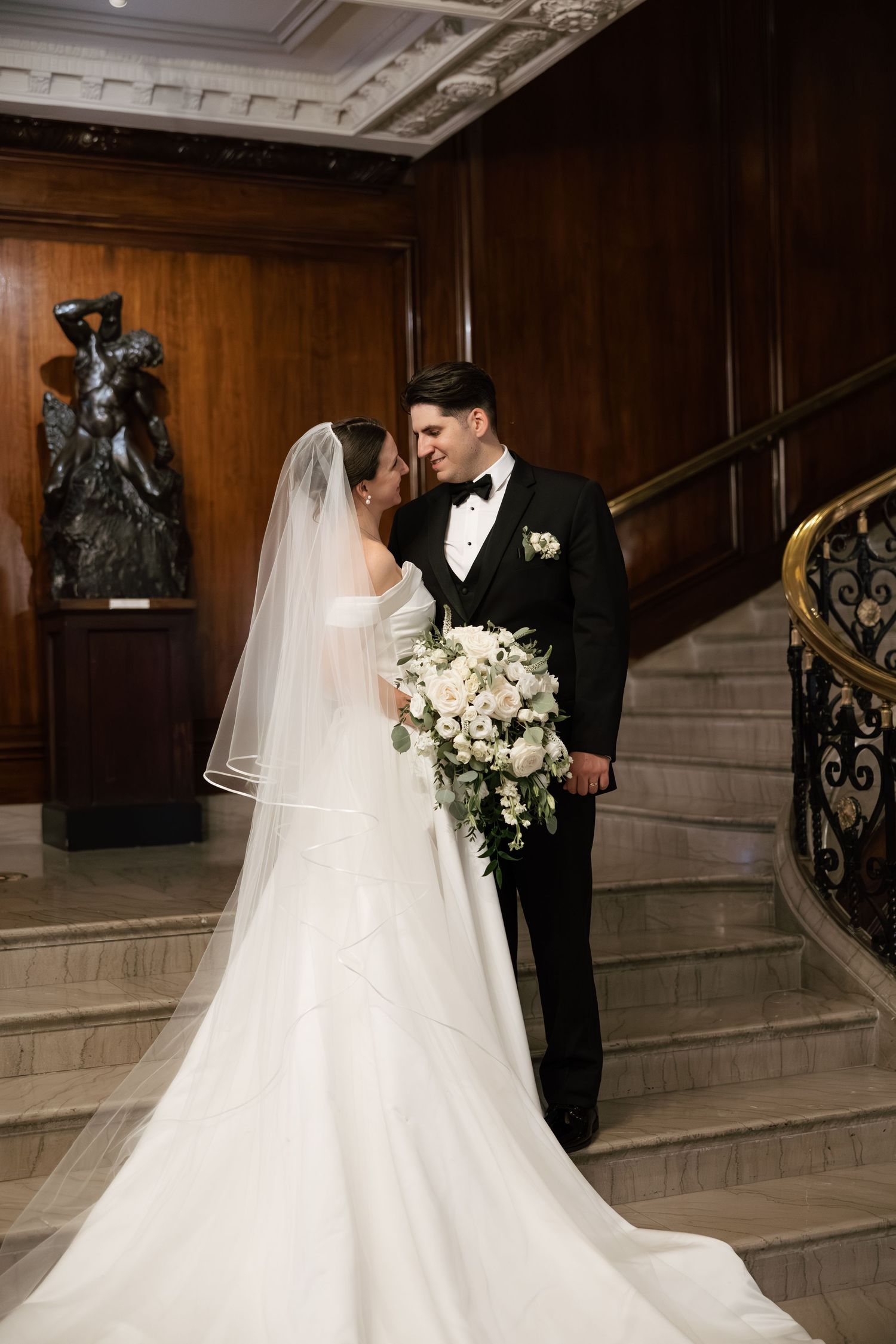 Elegant wedding couple shares intimate moment on grand staircase with ornate wood paneling and marble steps.