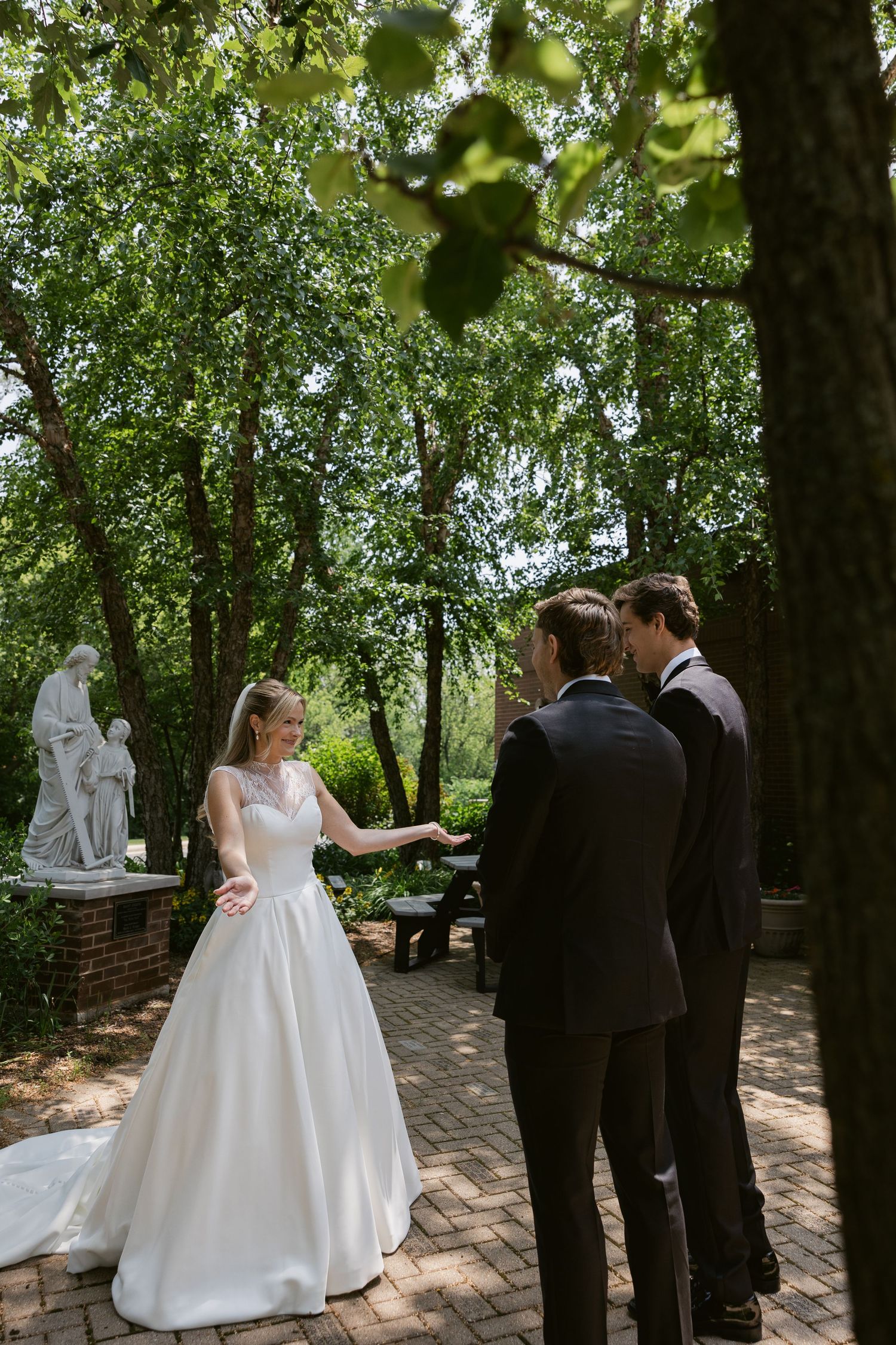 Wedding party gathers on a shaded brick path under trees for pre-ceremony photos.