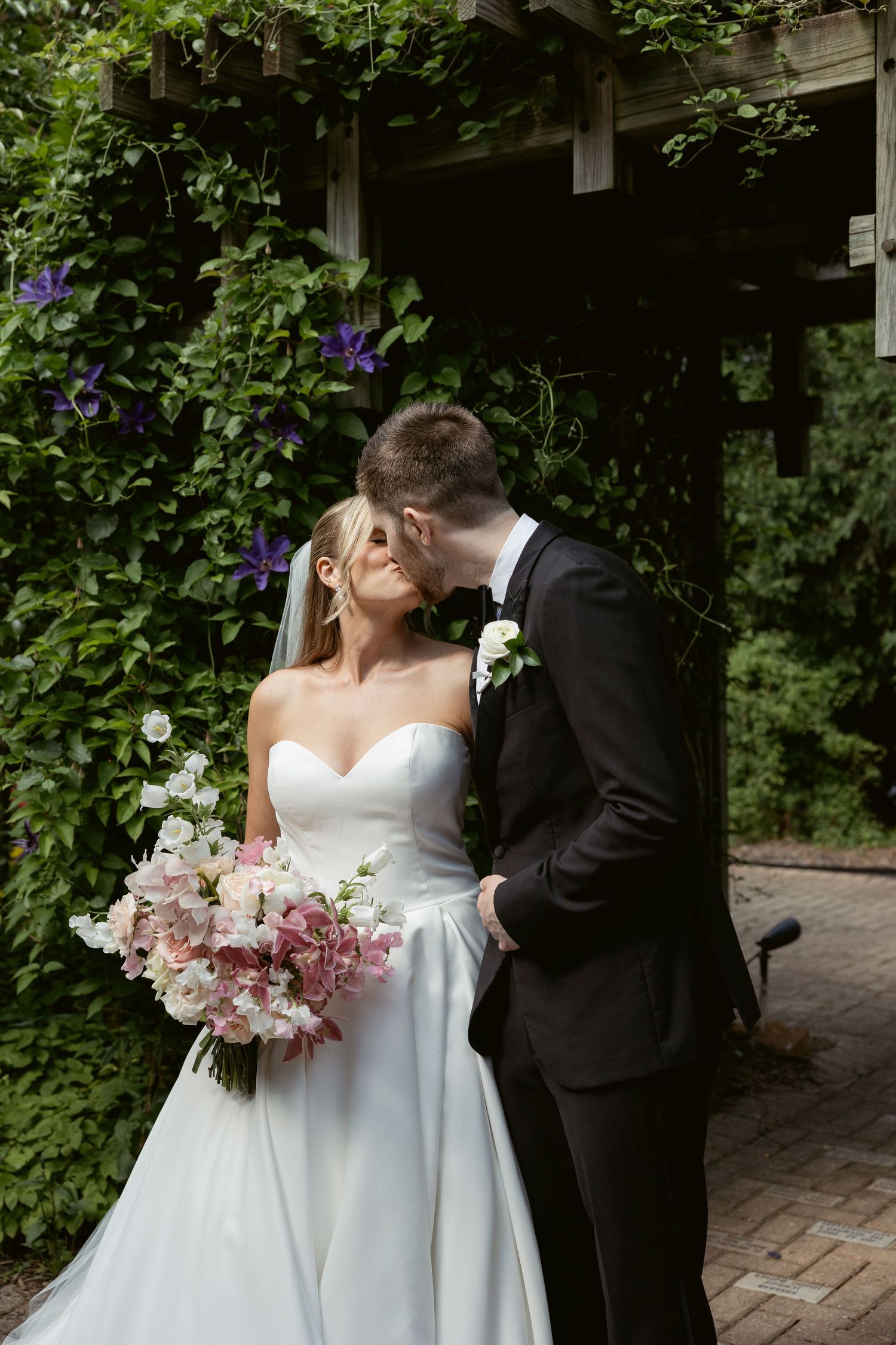 Romantic wedding couple embraces under garden archway with purple flowers and greenery.