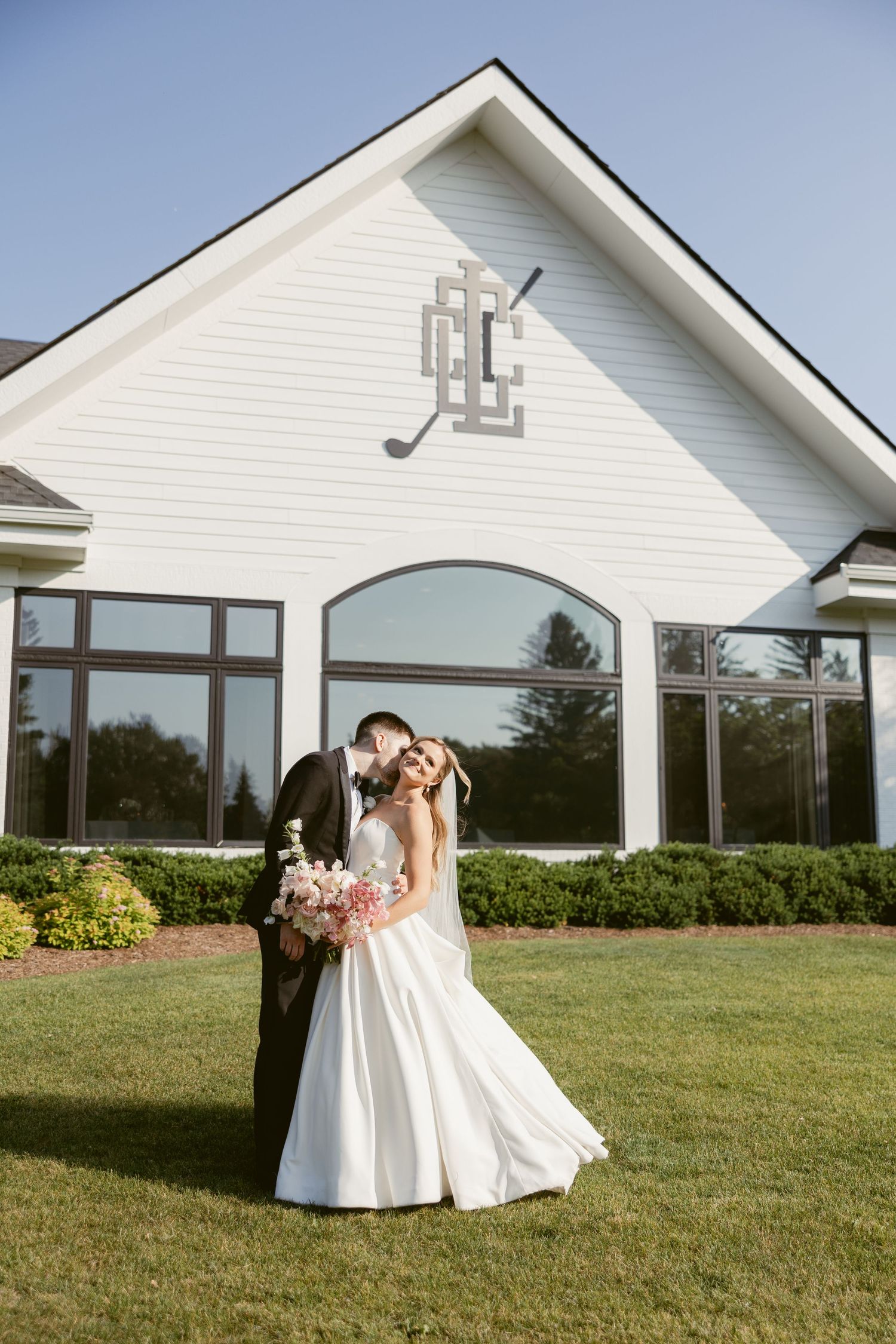 Wedding couple poses outside white clubhouse building with golf club emblem.