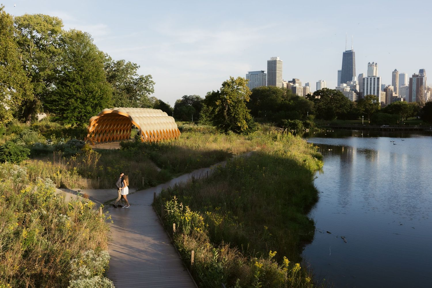 People walk along a lakeside path with the Chicago skyline visible in the distance on a sunny summer afternoon.