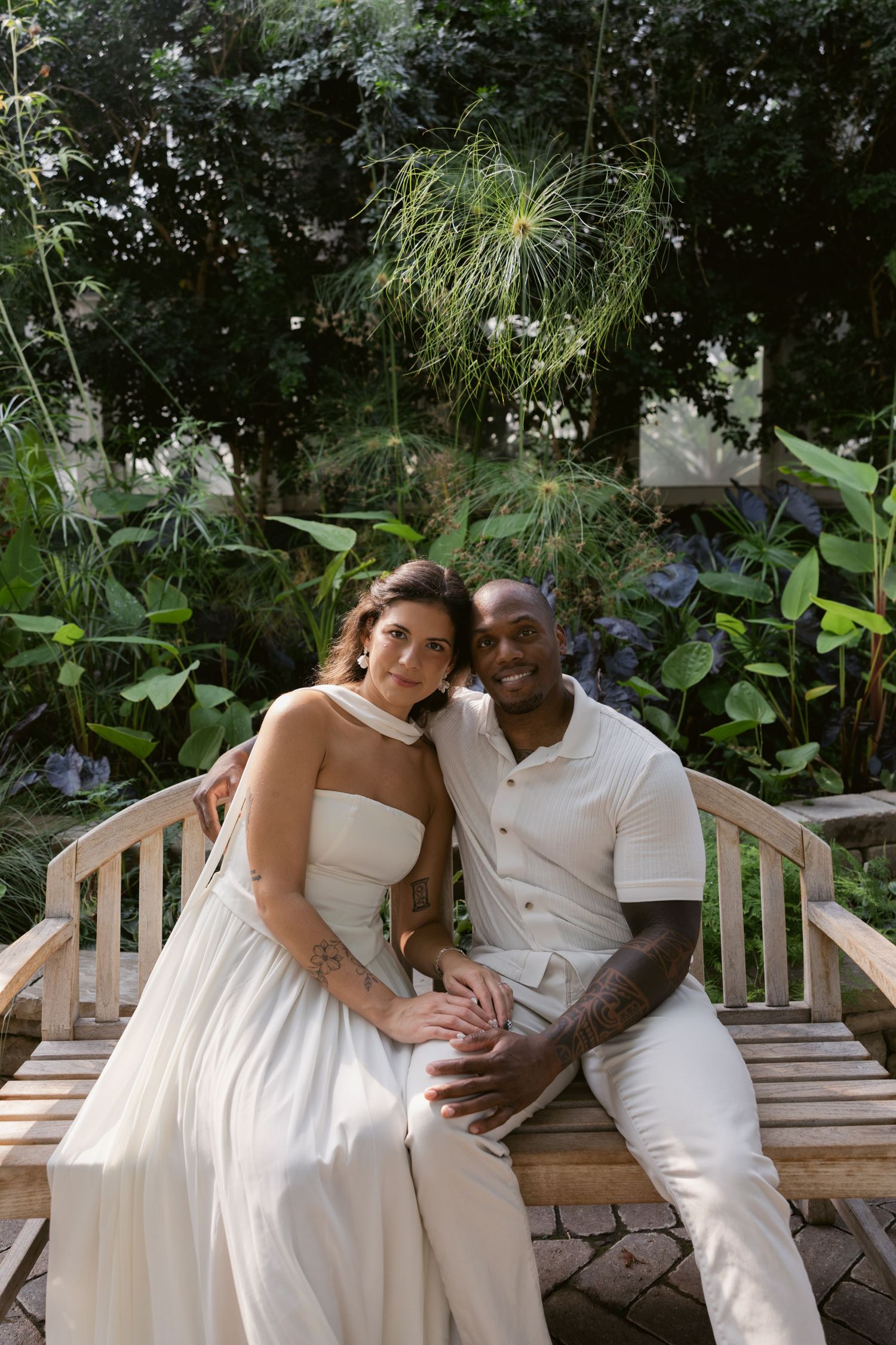 A romantic couple in white outfits sits together on a wooden bench in a lush garden setting.