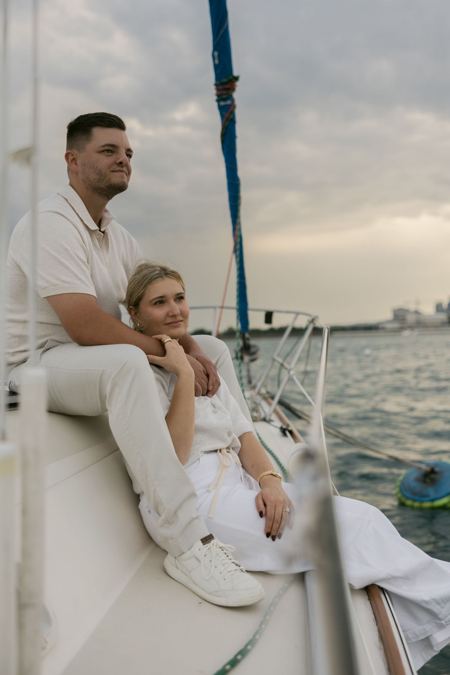 Couple in white outfits enjoying a peaceful moment on a sailboat with city skyline in background.