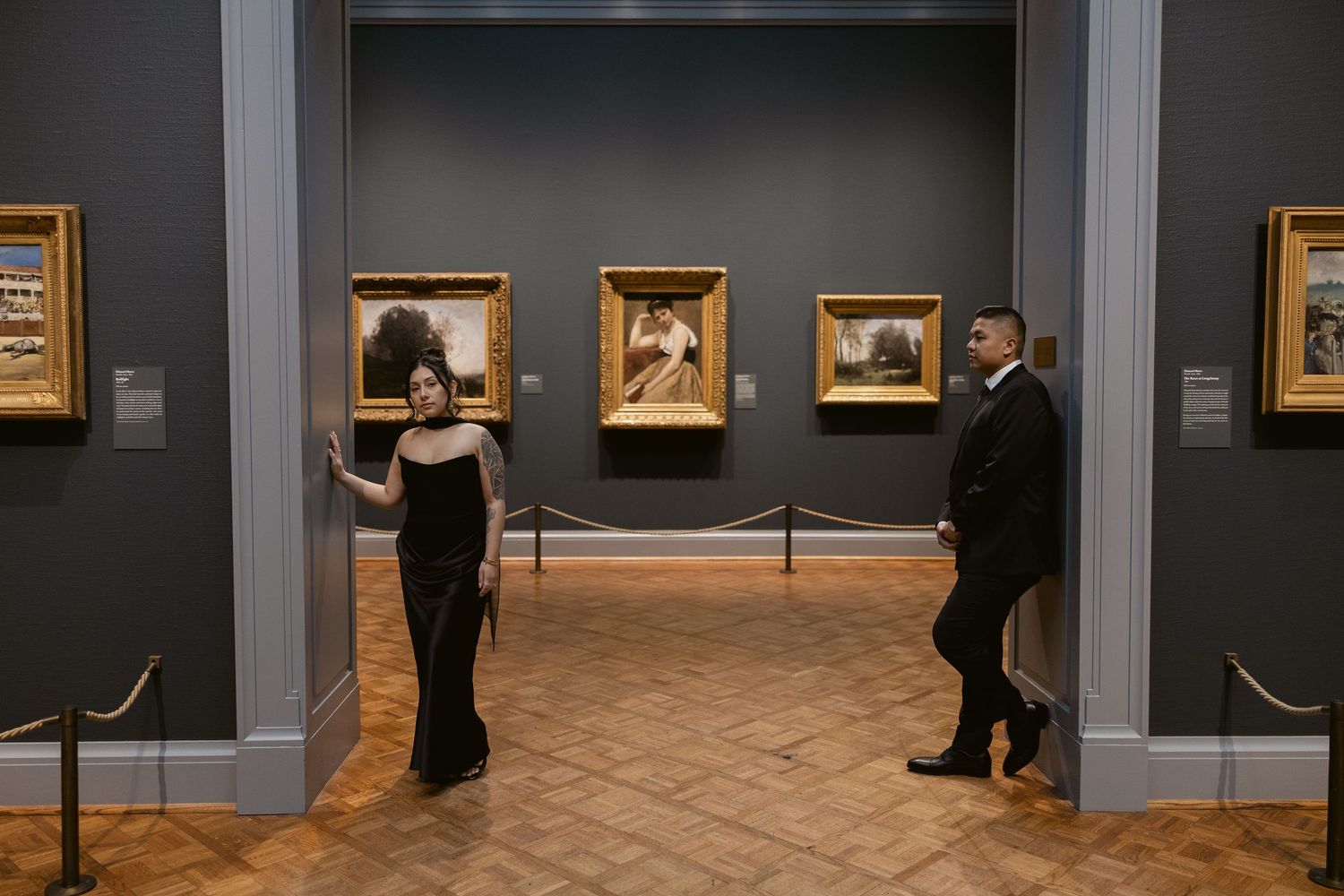 A couple in formal wear walks through an art gallery with paintings on dark walls.