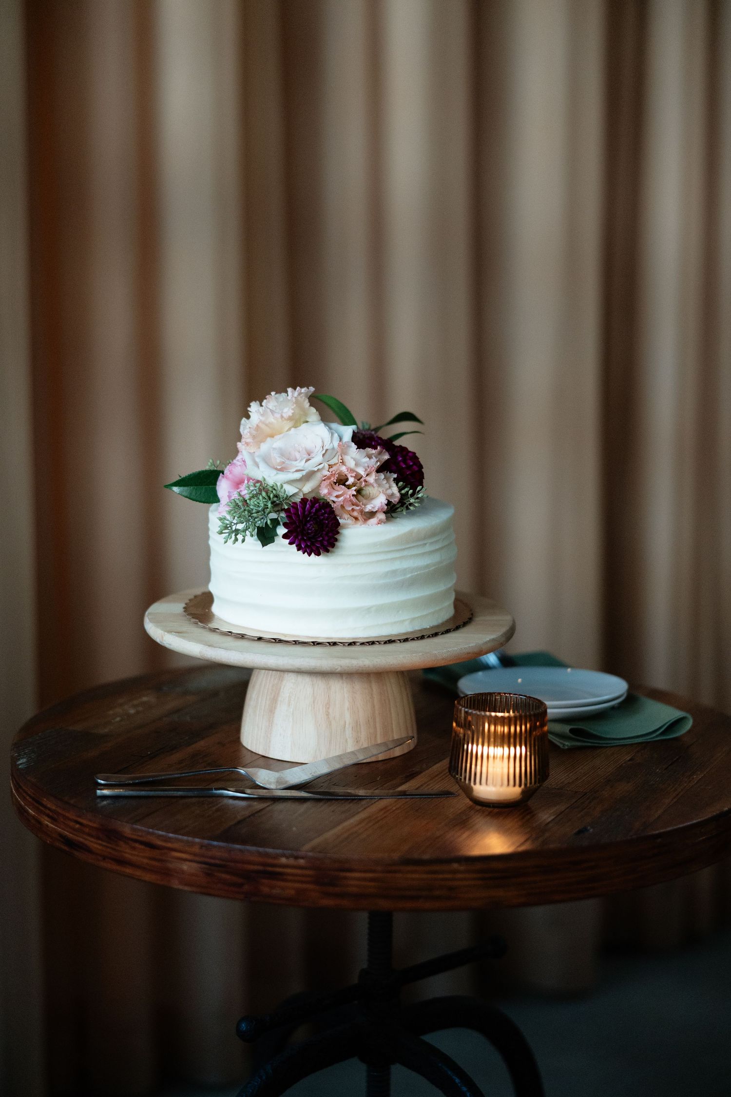 Three white wedding cakes decorated with flowers sit on wooden tables with ambient candlelight and draped fabric backdrop.