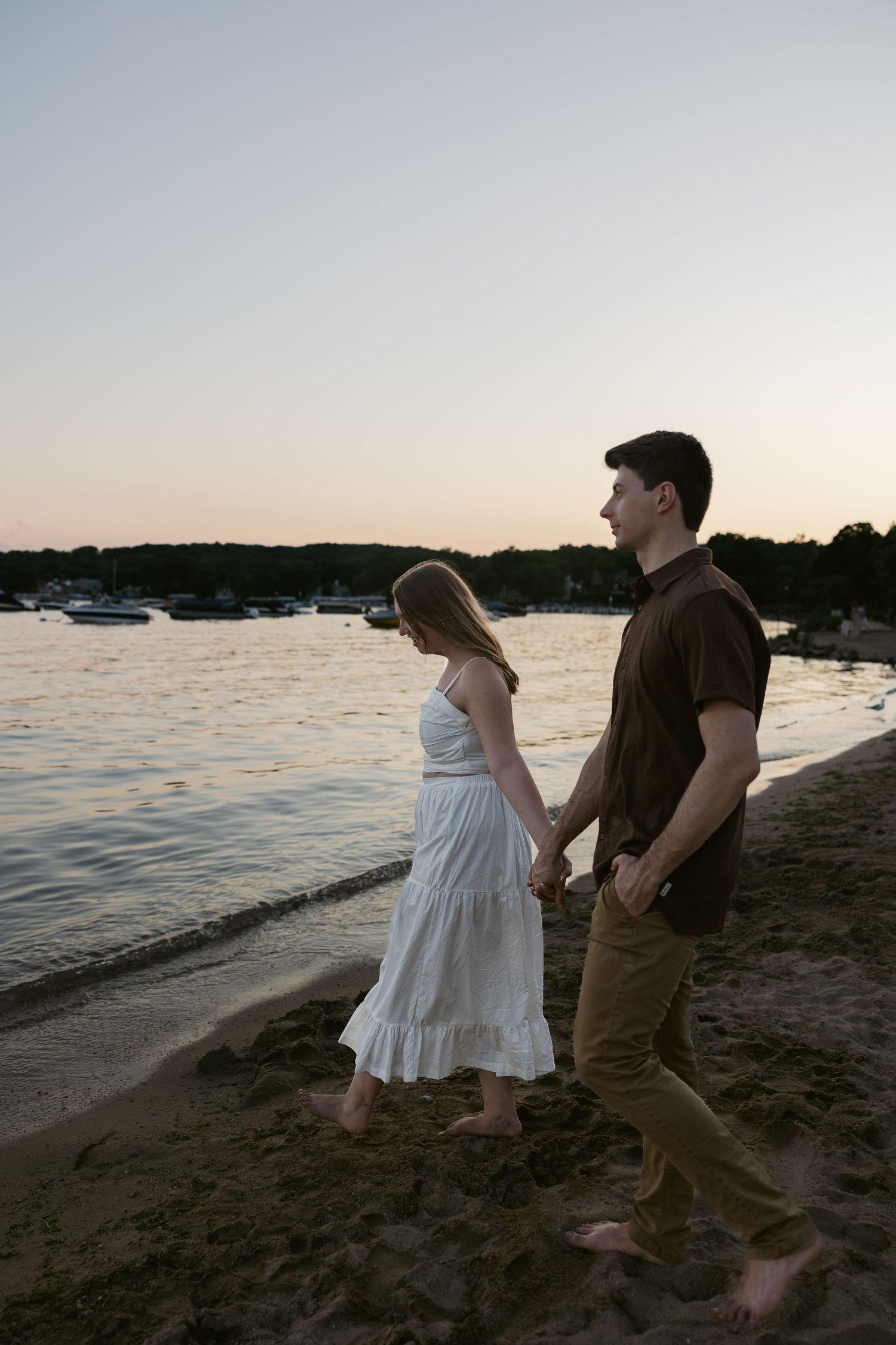 Couple dancing playfully along a beach shoreline at sunset with boats visible in the distance.