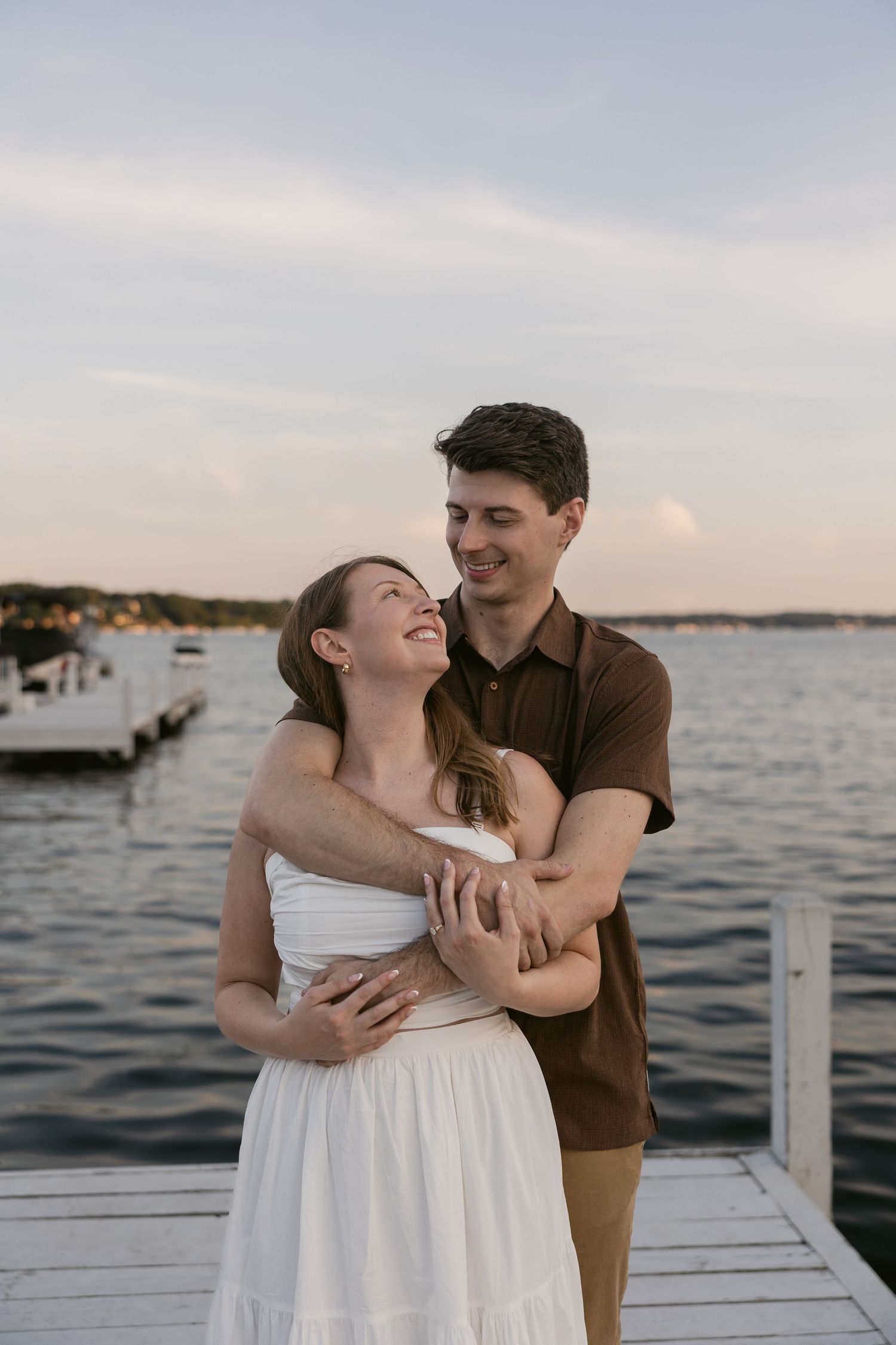 A romantic couple embraces on a dock at sunset with boats and water in the background.