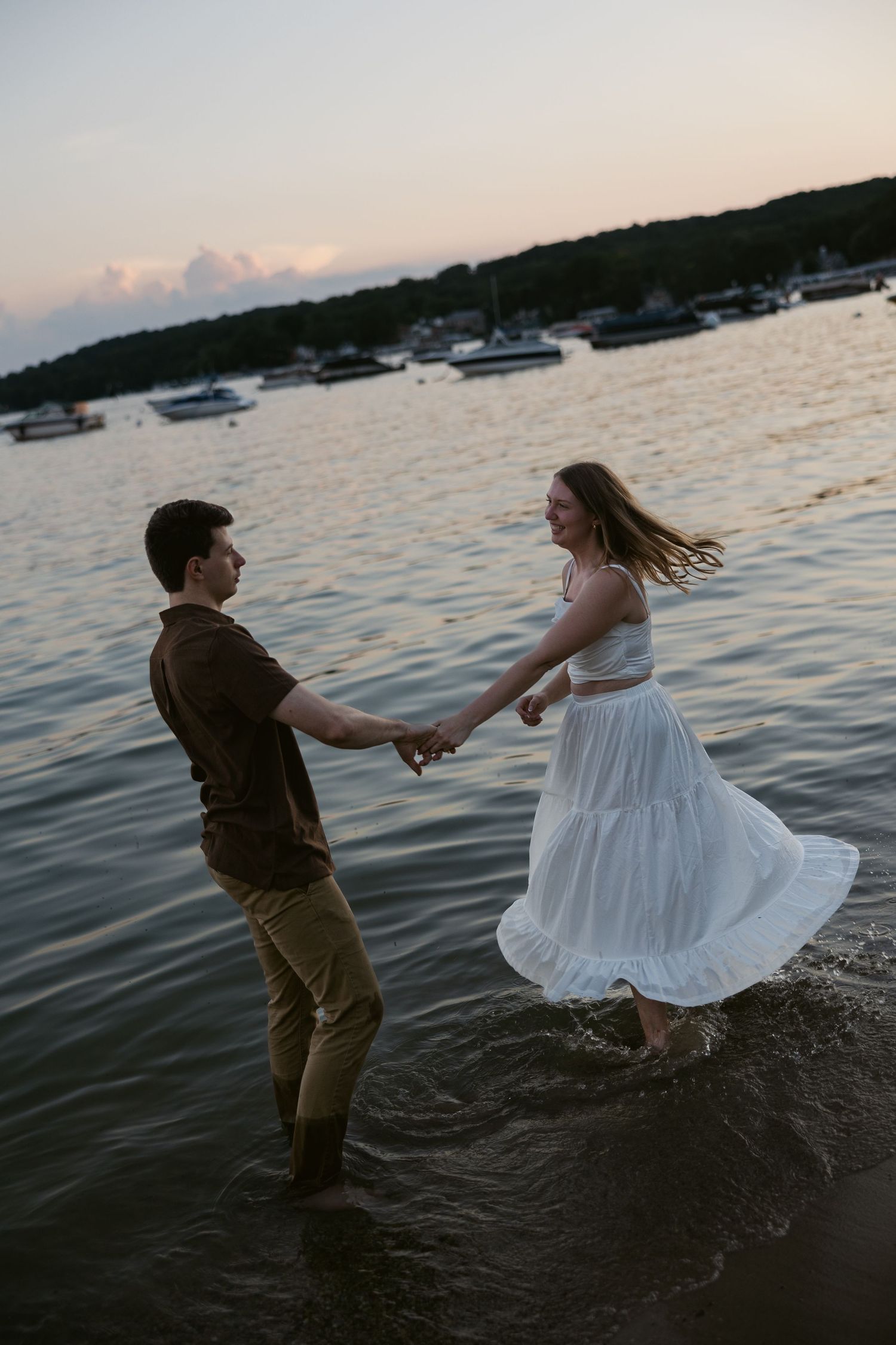 A couple dances playfully at the water's edge during sunset with boats in the background.