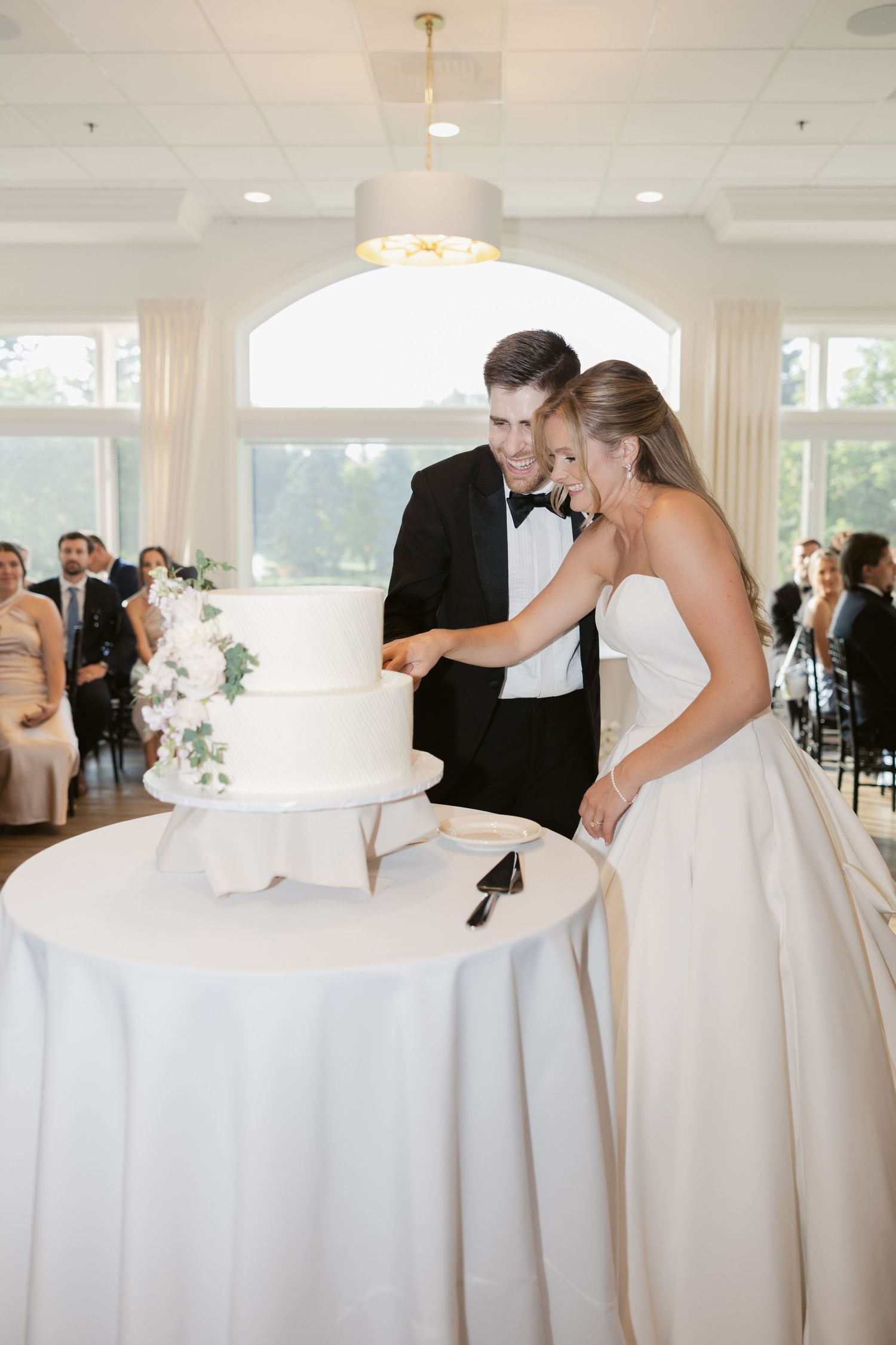 A tiered white wedding cake sits on an elegant table at a reception while guests celebrate in the background.
