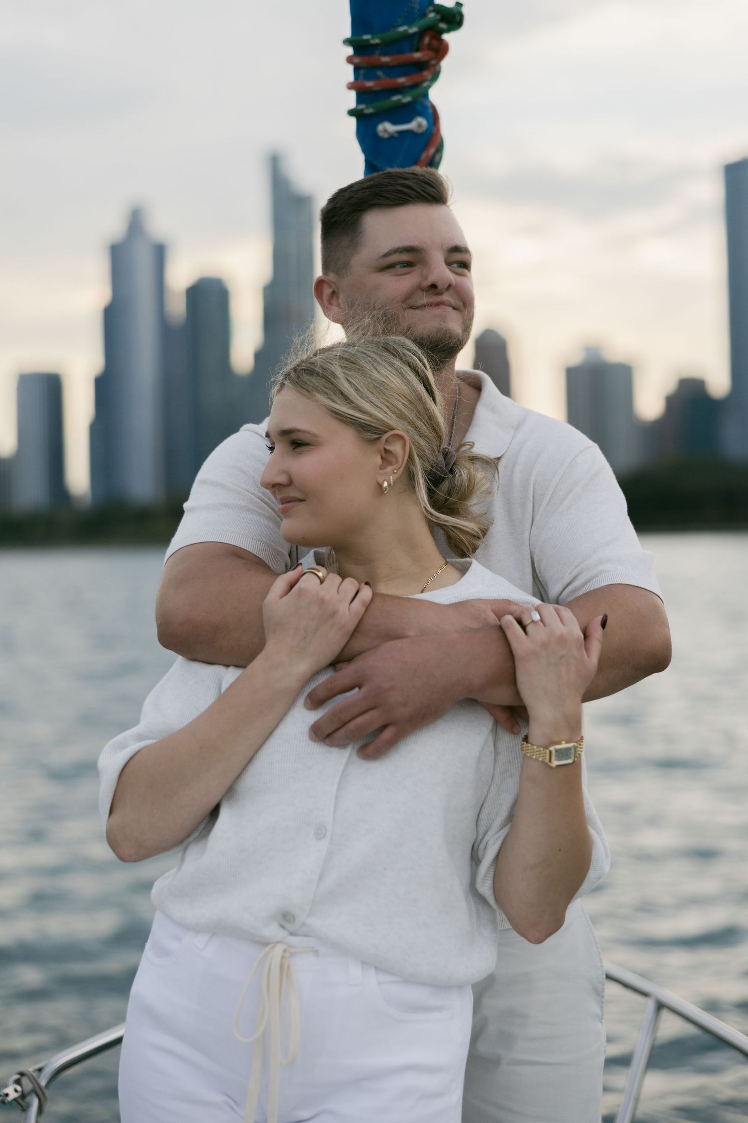 A couple dressed in all white embraces on a sailboat with a cityscape visible across the water.