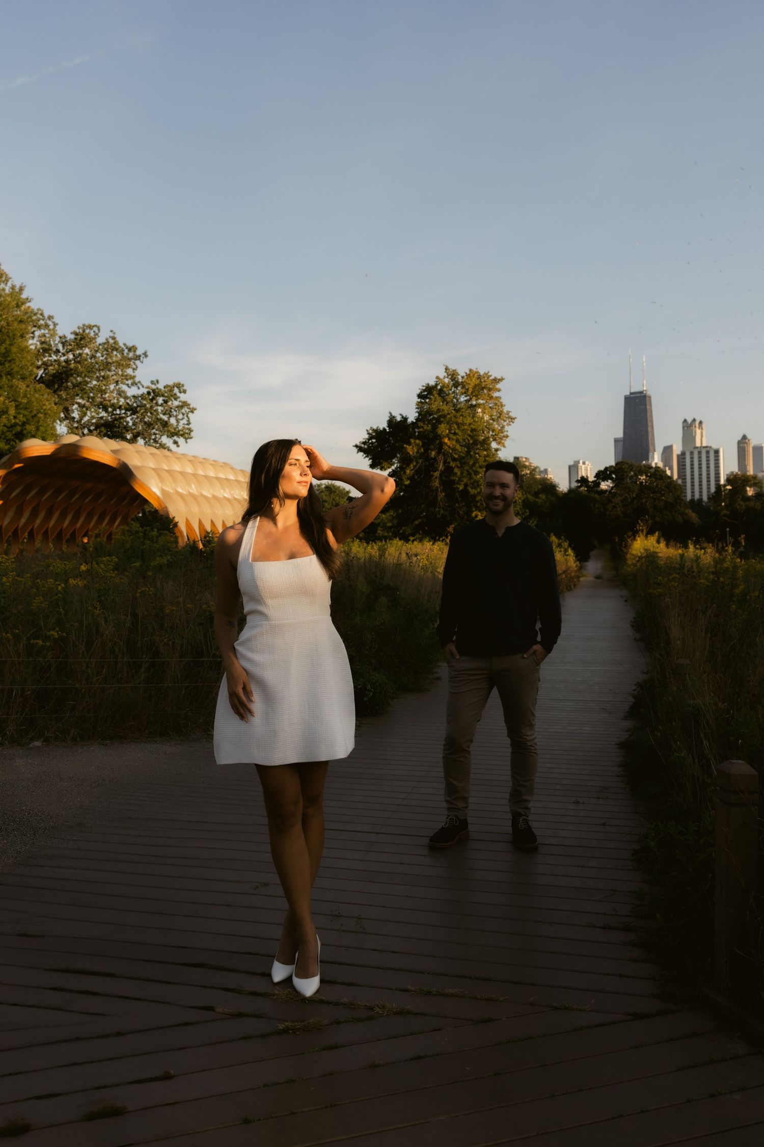A couple poses at sunset on a wooden deck with a city skyline visible in the distance.