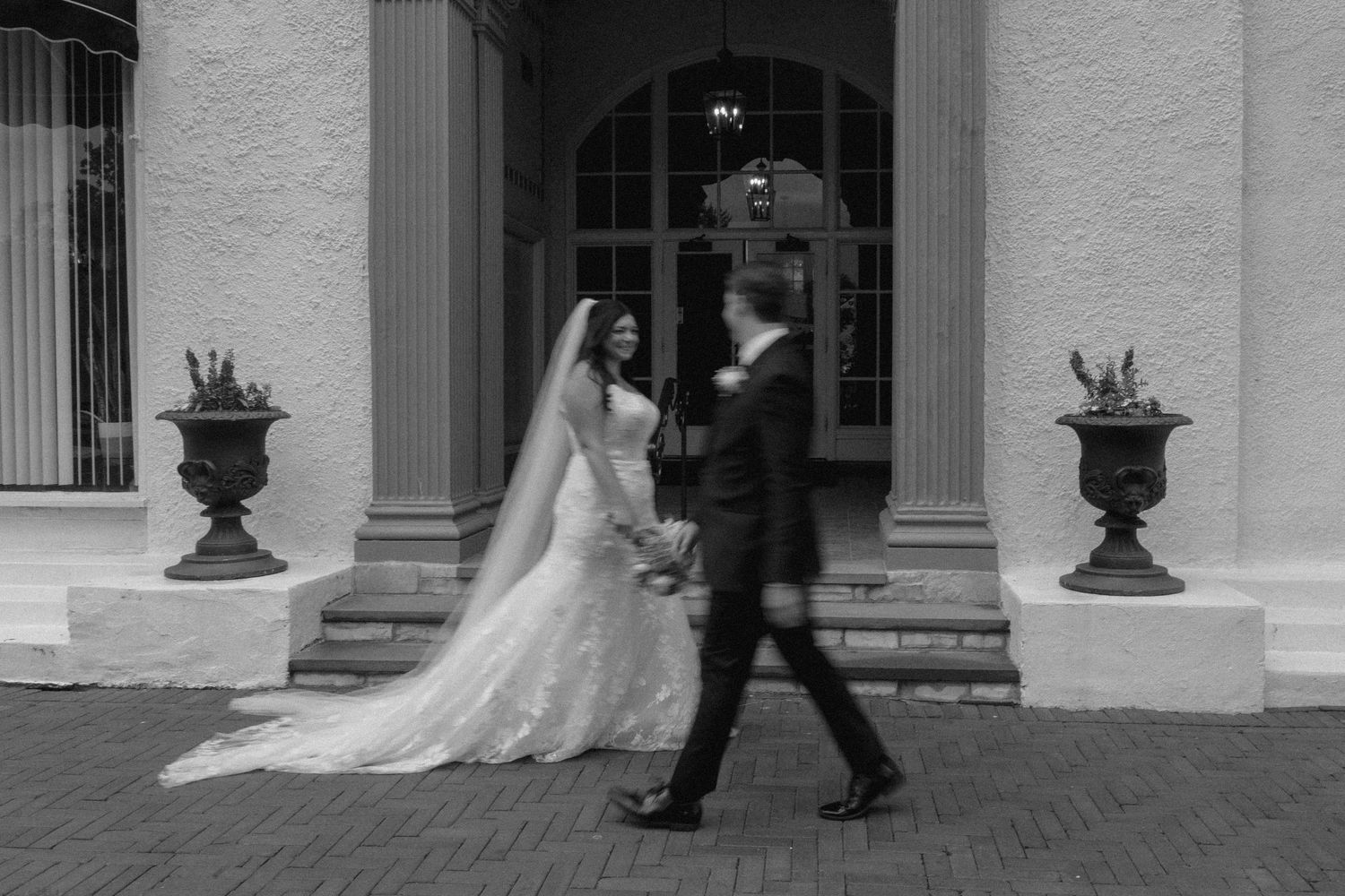 Black and white photo of a bride and groom walking between classical columns with decorative urns at a wedding venue.