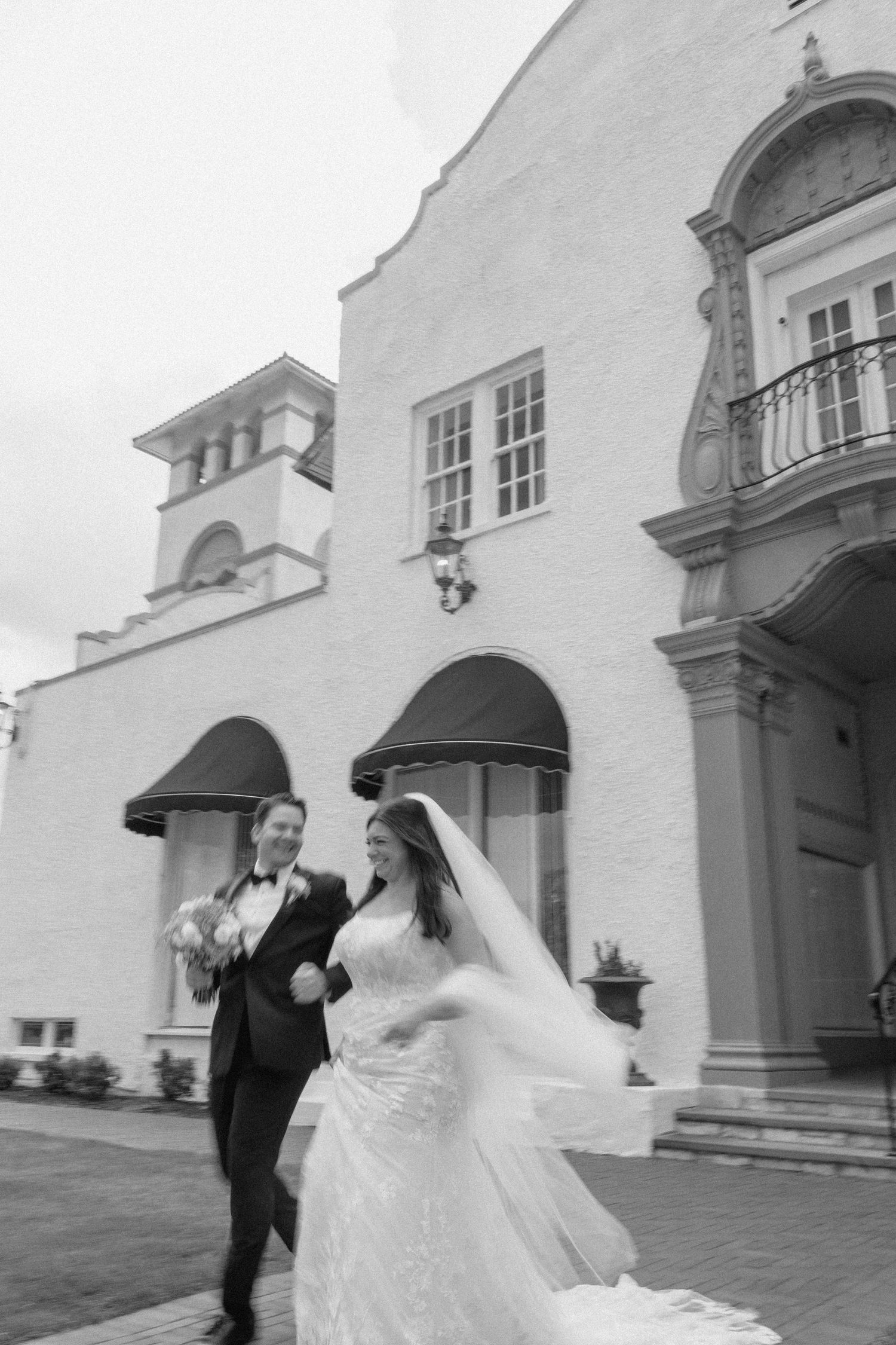 A couple in wedding attire walking outside a white Spanish colonial style building with arched doorways and balconies.