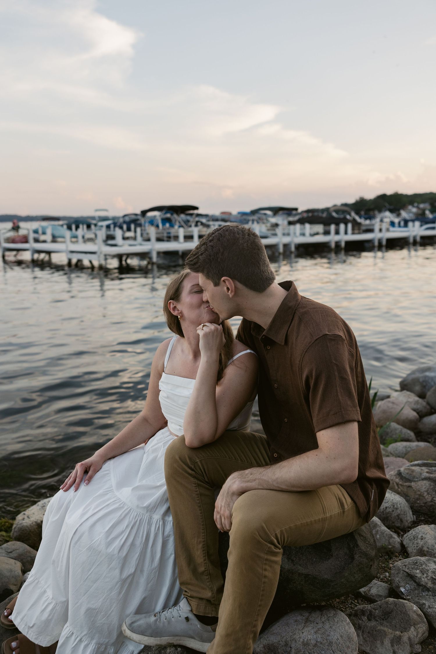 Intimate moment shared between couple on rocky shore with boats in background at sunset.
