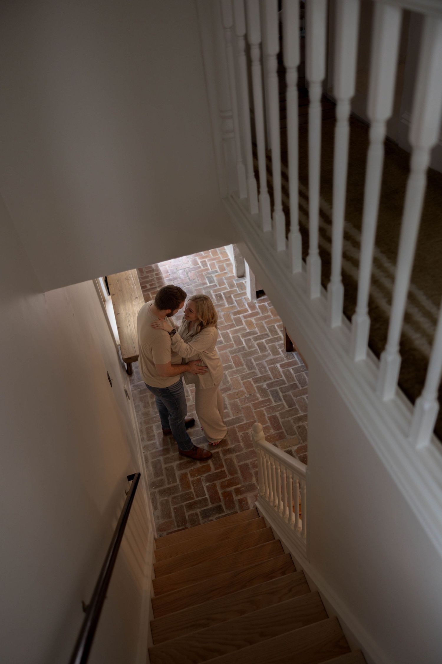 A downward view of wooden stairs with a white banister shows figures in a curved herringbone-patterned hallway below.