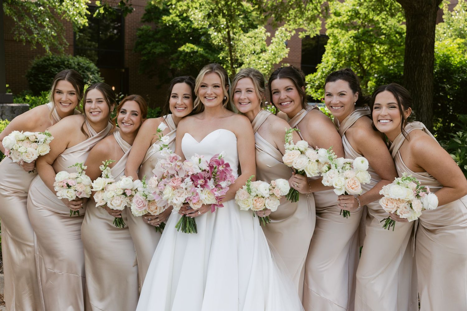 Wedding party members in neutral dresses posing with floral bouquets in a garden setting.