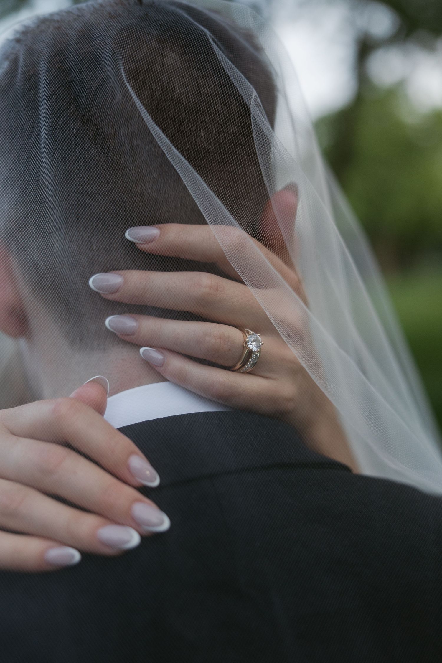 Close-up of bride's hands with wedding ring embracing groom's shoulder through delicate veil.