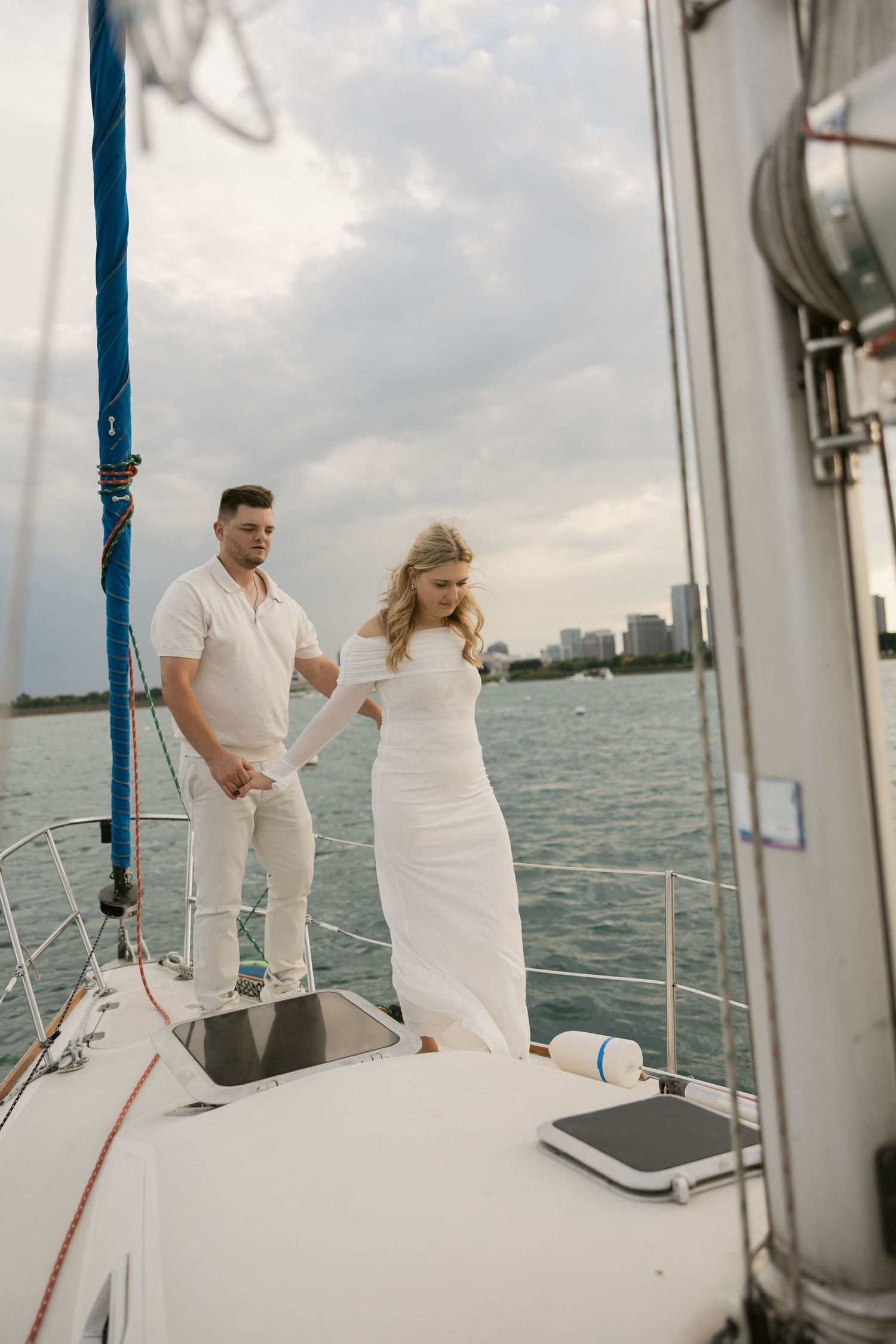 Couple dressed in white aboard a sailboat with city skyline visible in the distance.