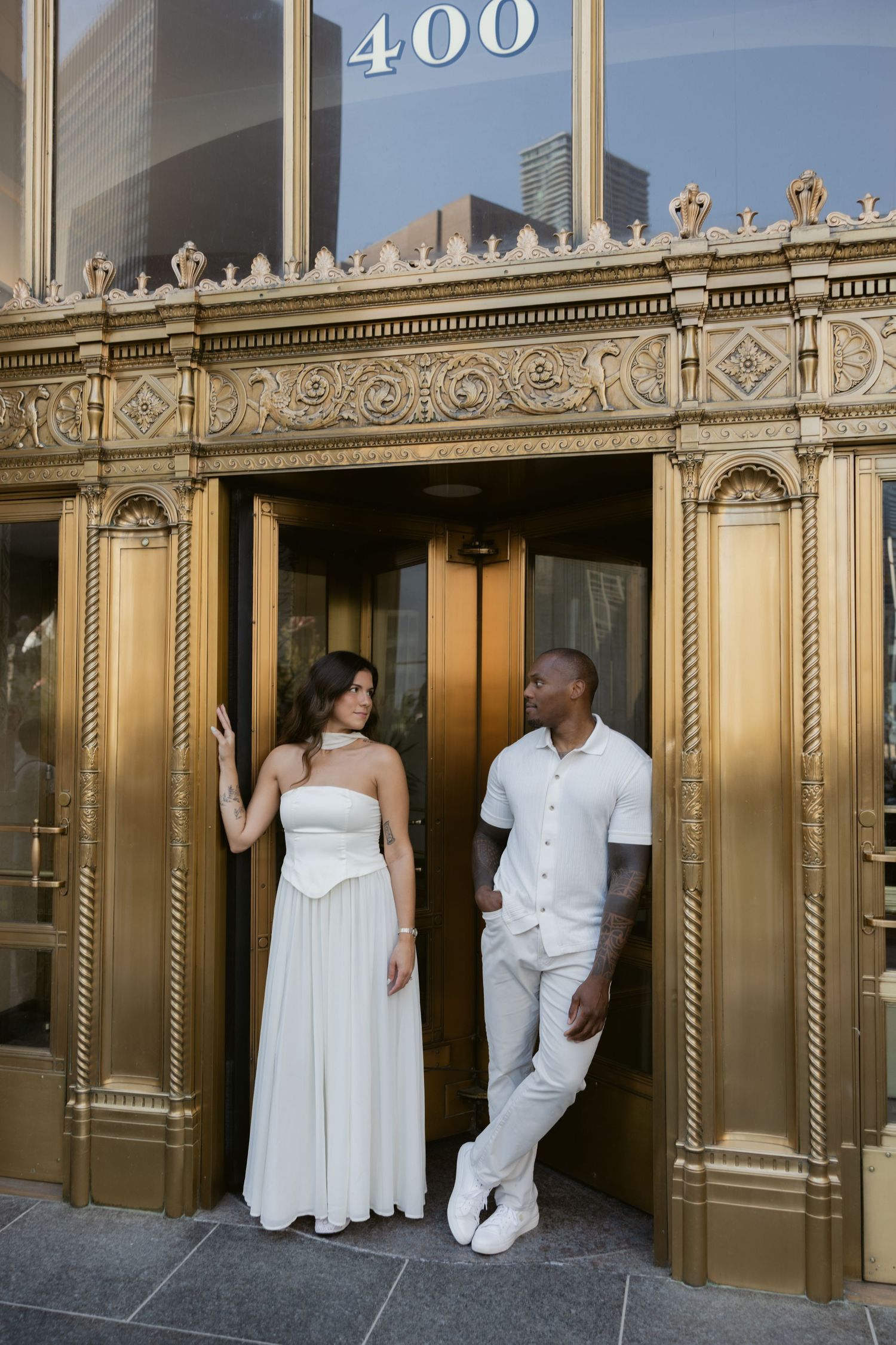 Couple in white outfits posing against ornate gold architectural details of building entrance.