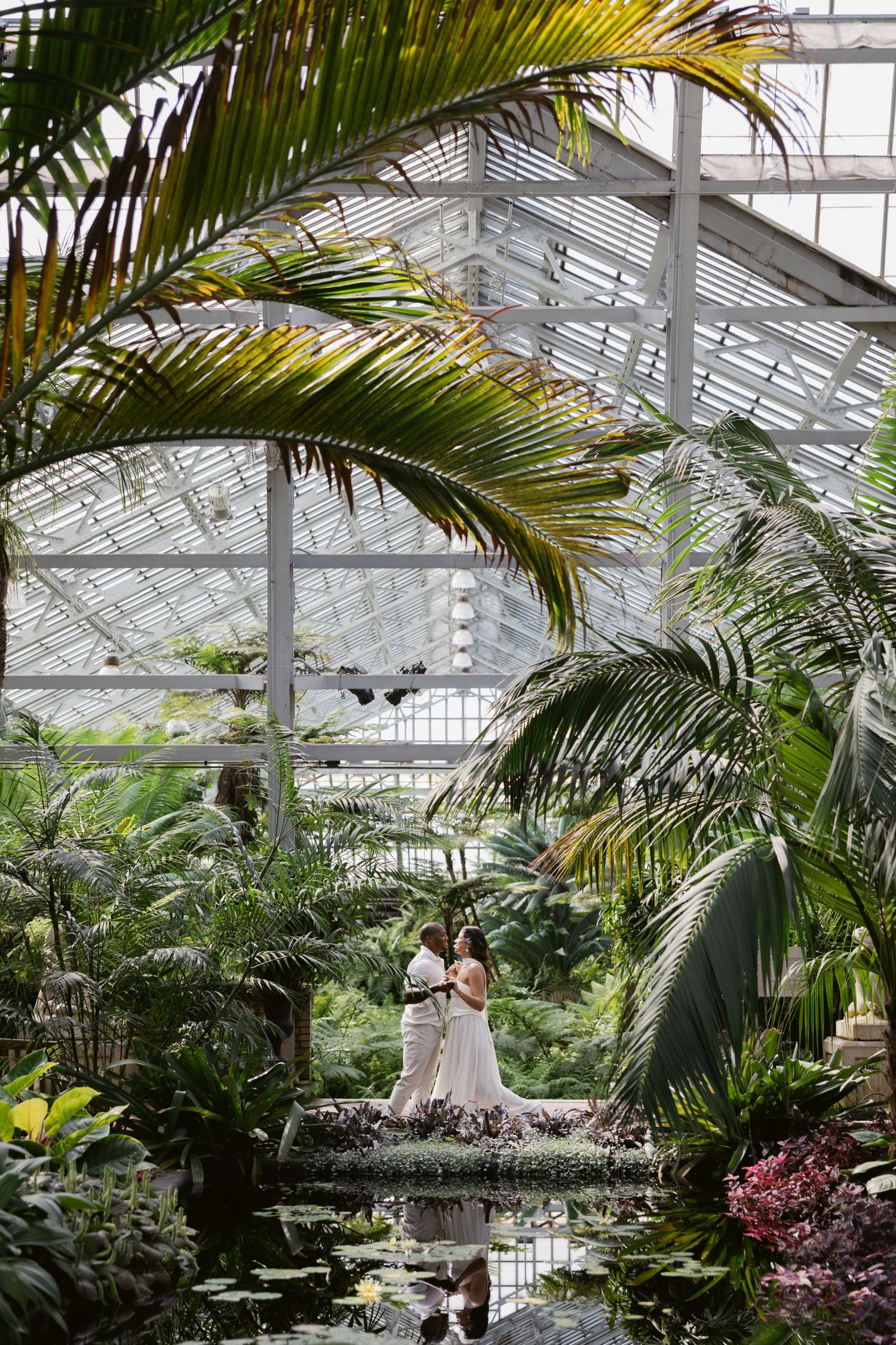 A romantic wedding photoshoot in a lush greenhouse conservatory filled with tropical palm fronds and natural light.