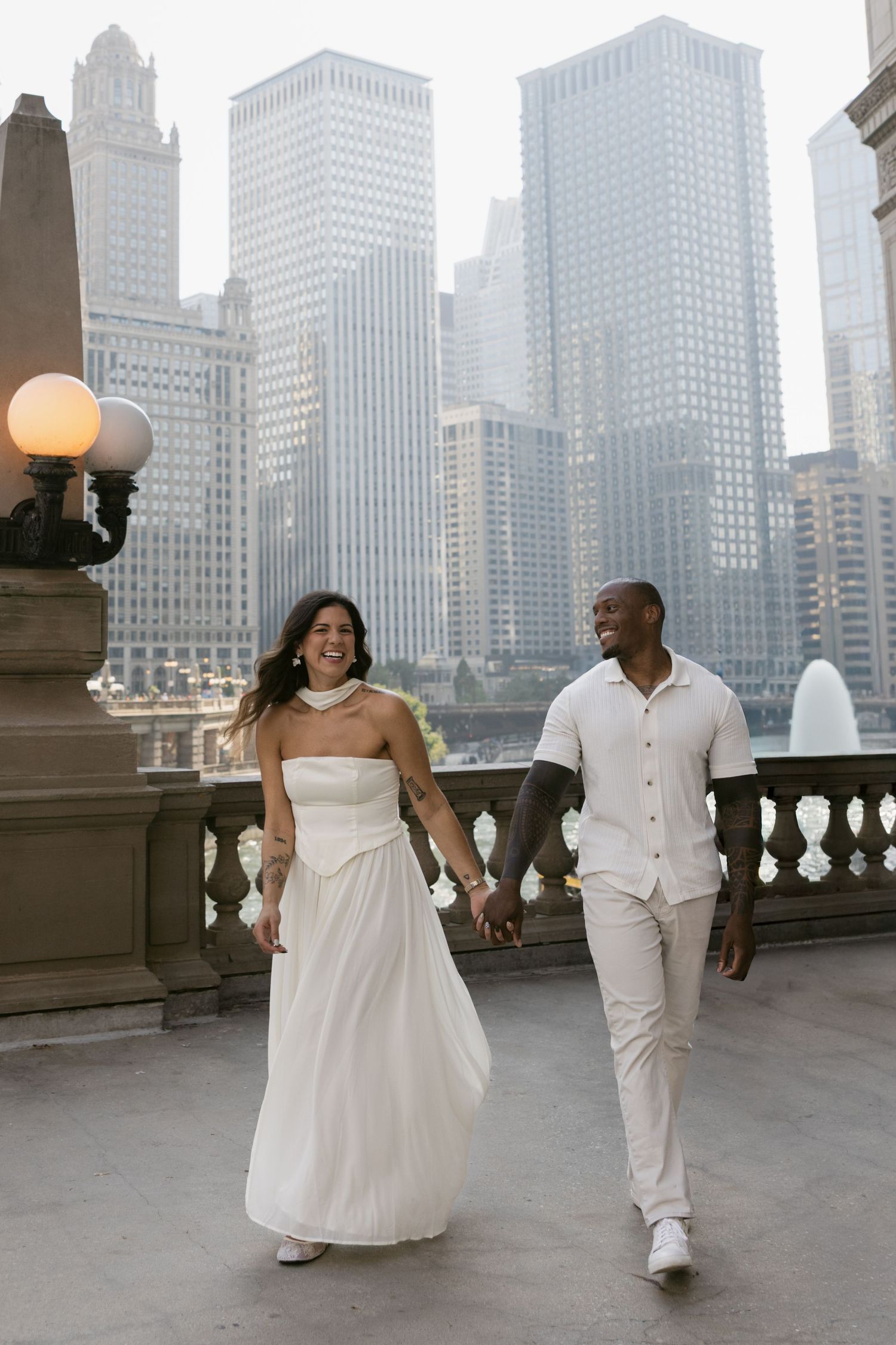 A couple in white outfits walks hand in hand on a rooftop terrace with Chicago's dramatic skyline as backdrop.