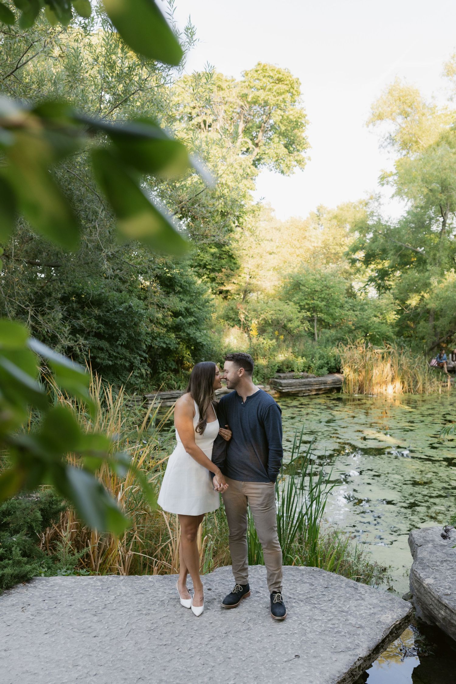 A romantic couple poses by a serene lily pond surrounded by lush greenery on a sunny day.