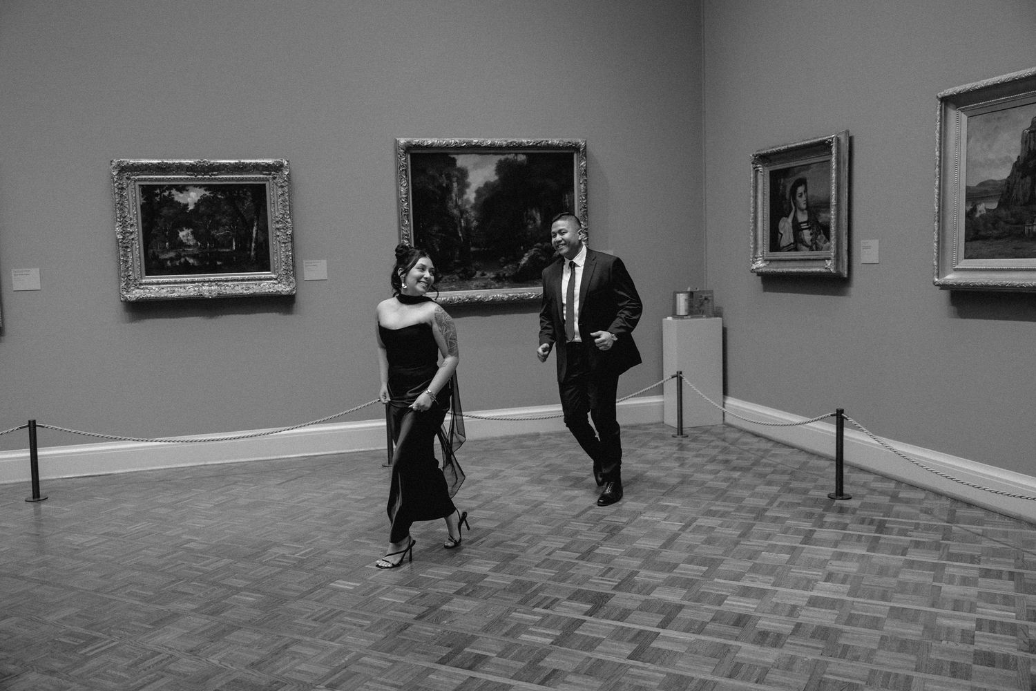 A couple explores an art museum gallery with framed paintings on grey walls and parquet flooring.