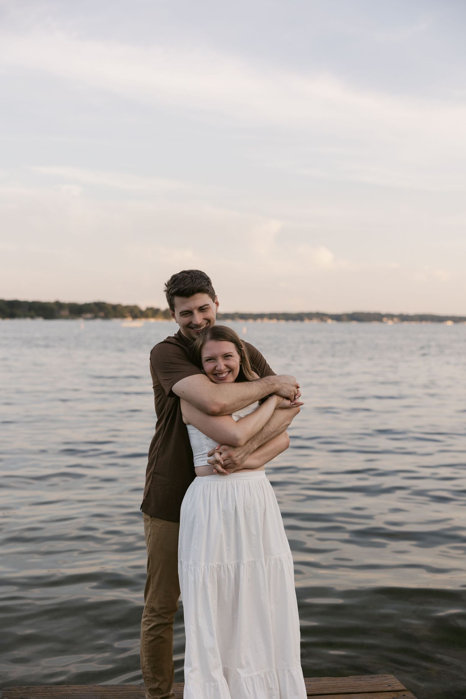 A romantic sunset photoshoot on a dock by the water with a couple embracing in casual summer attire.