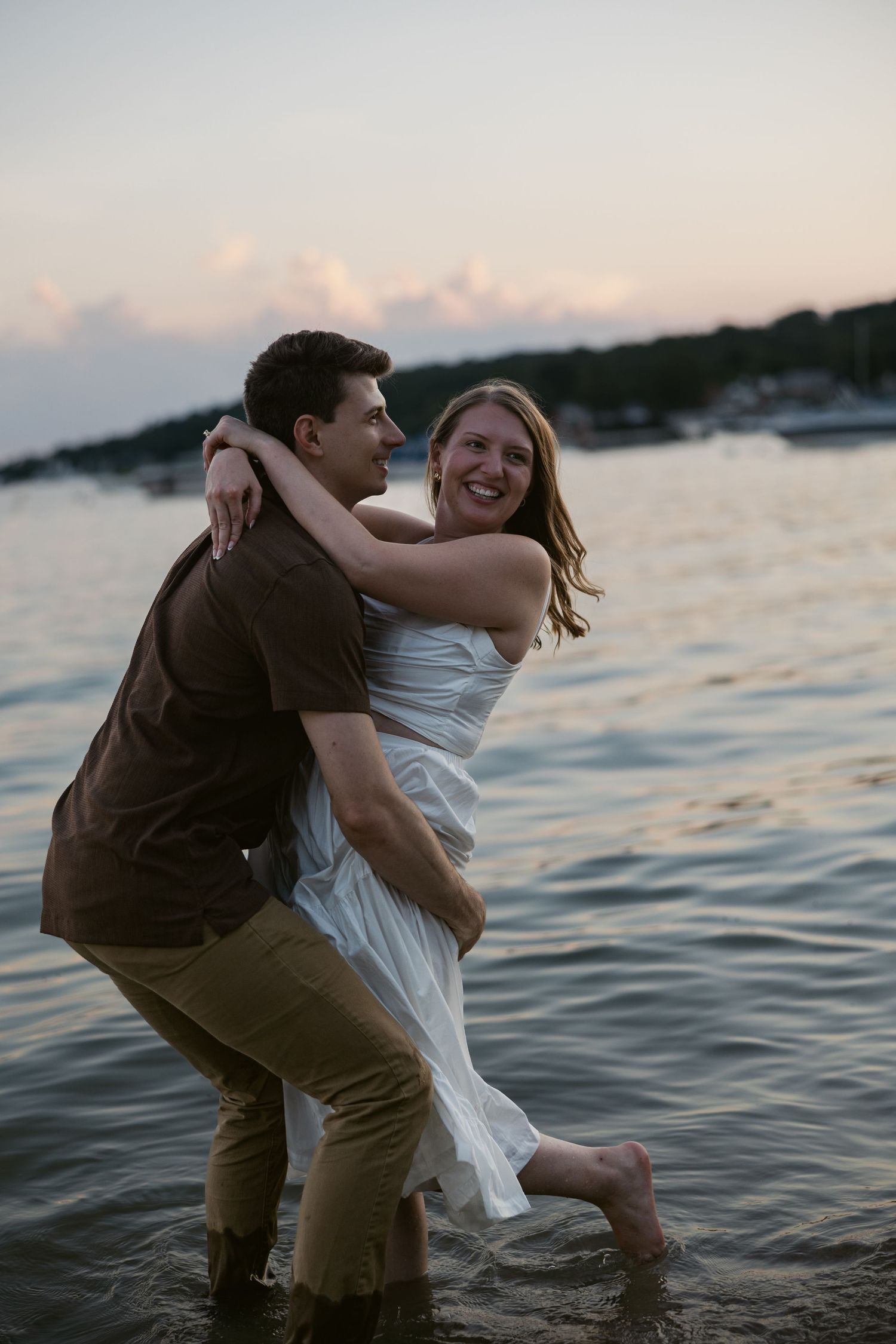 A playful couple splashes in the water at sunset near a marina.