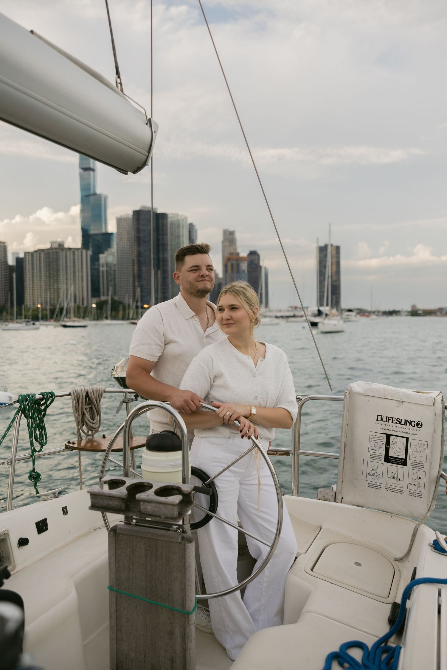Two people in matching white outfits navigate a sailboat with downtown Detroit visible across the water.