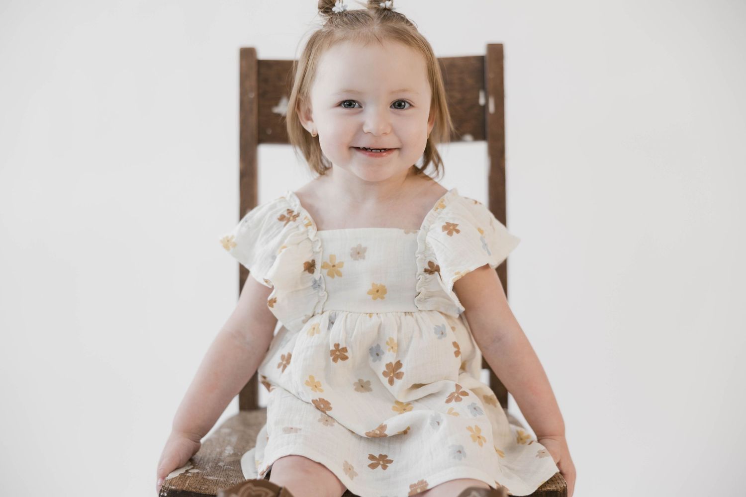 Toddler in white floral dress smiling while seated on wooden chair against white background.