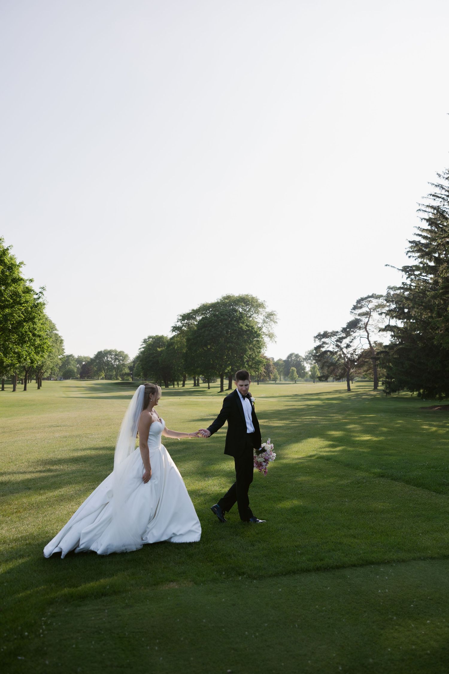 A couple walks hand in hand across a scenic golf course during their wedding photoshoot.