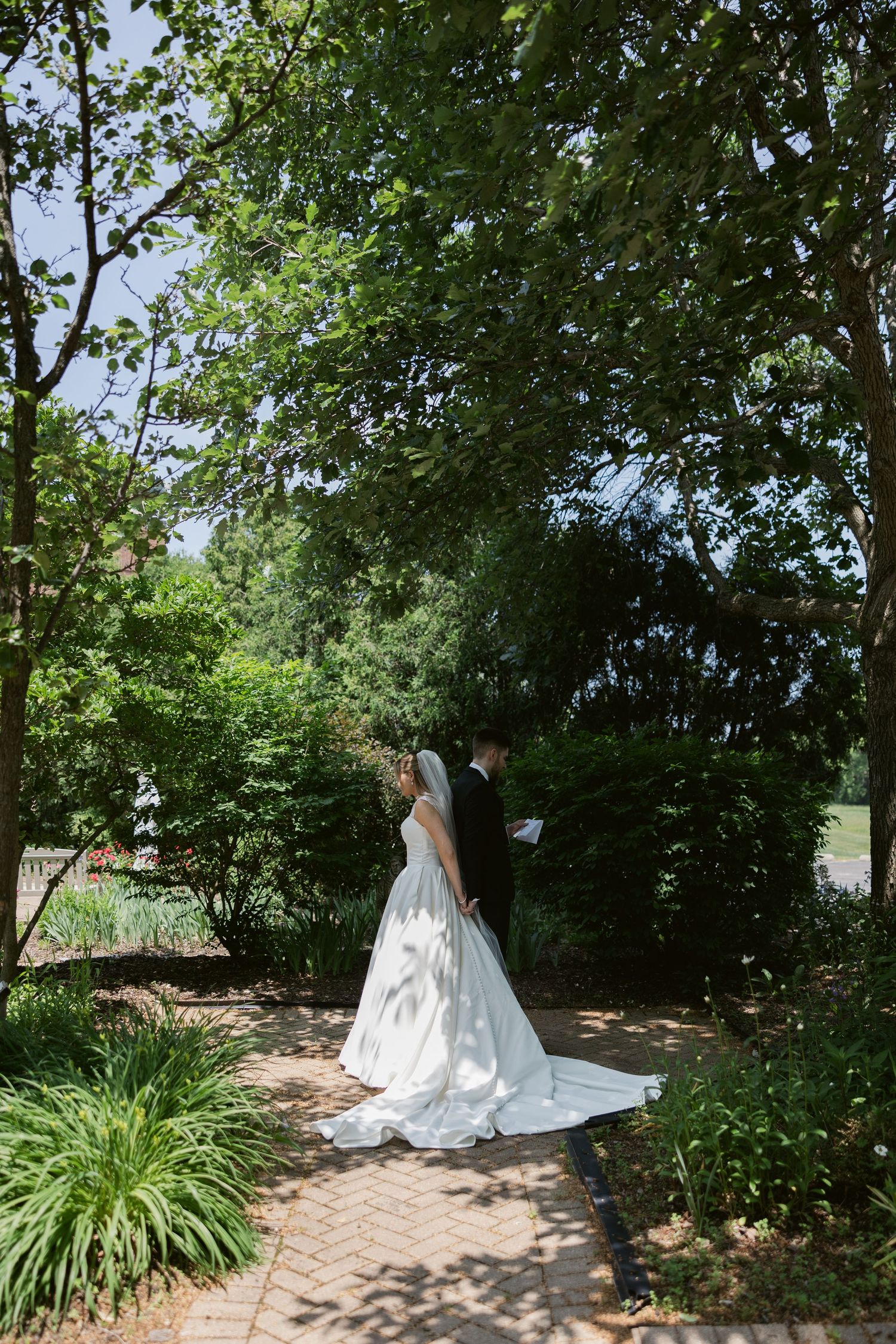A beautiful white wedding dress with a long train flows along a brick pathway surrounded by lush green gardens.
