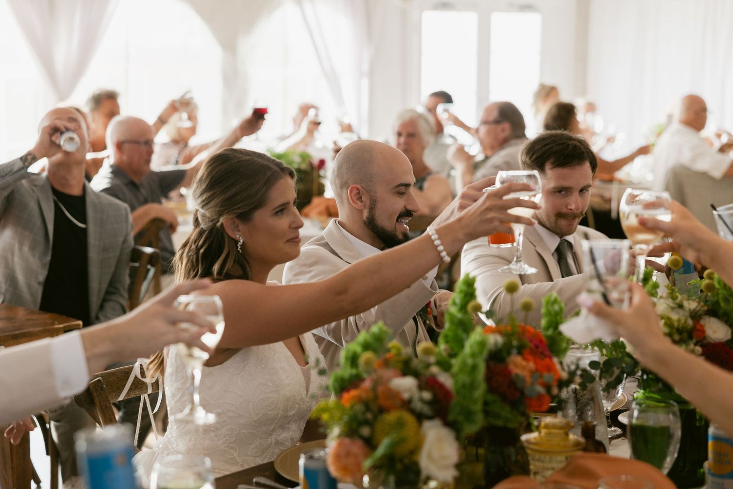 People raise their drinks in a festive toast at a celebratory gathering with flowers and natural lighting.