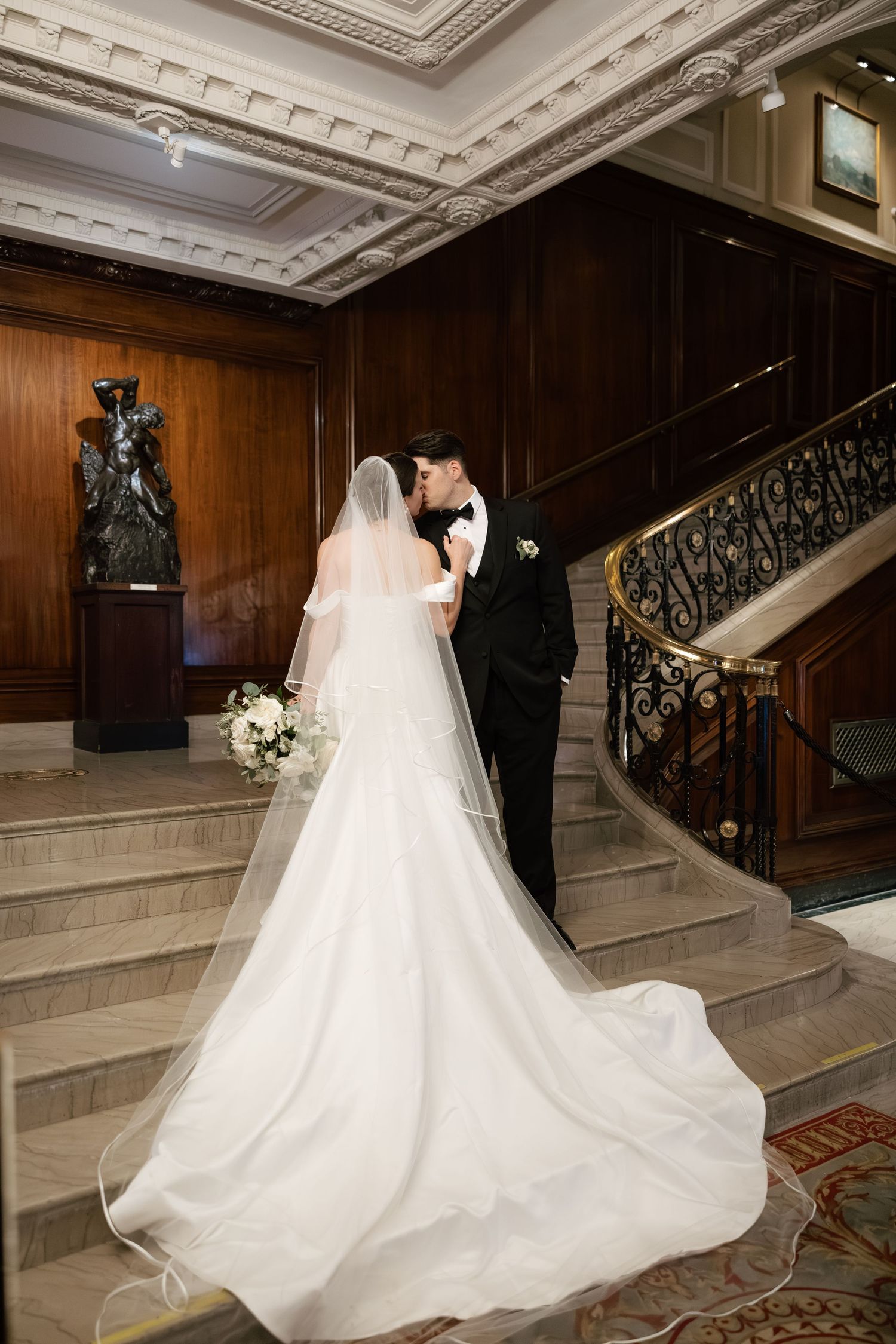 A couple in wedding attire pose on an elegant staircase with wood paneling and ornate ceiling details.
