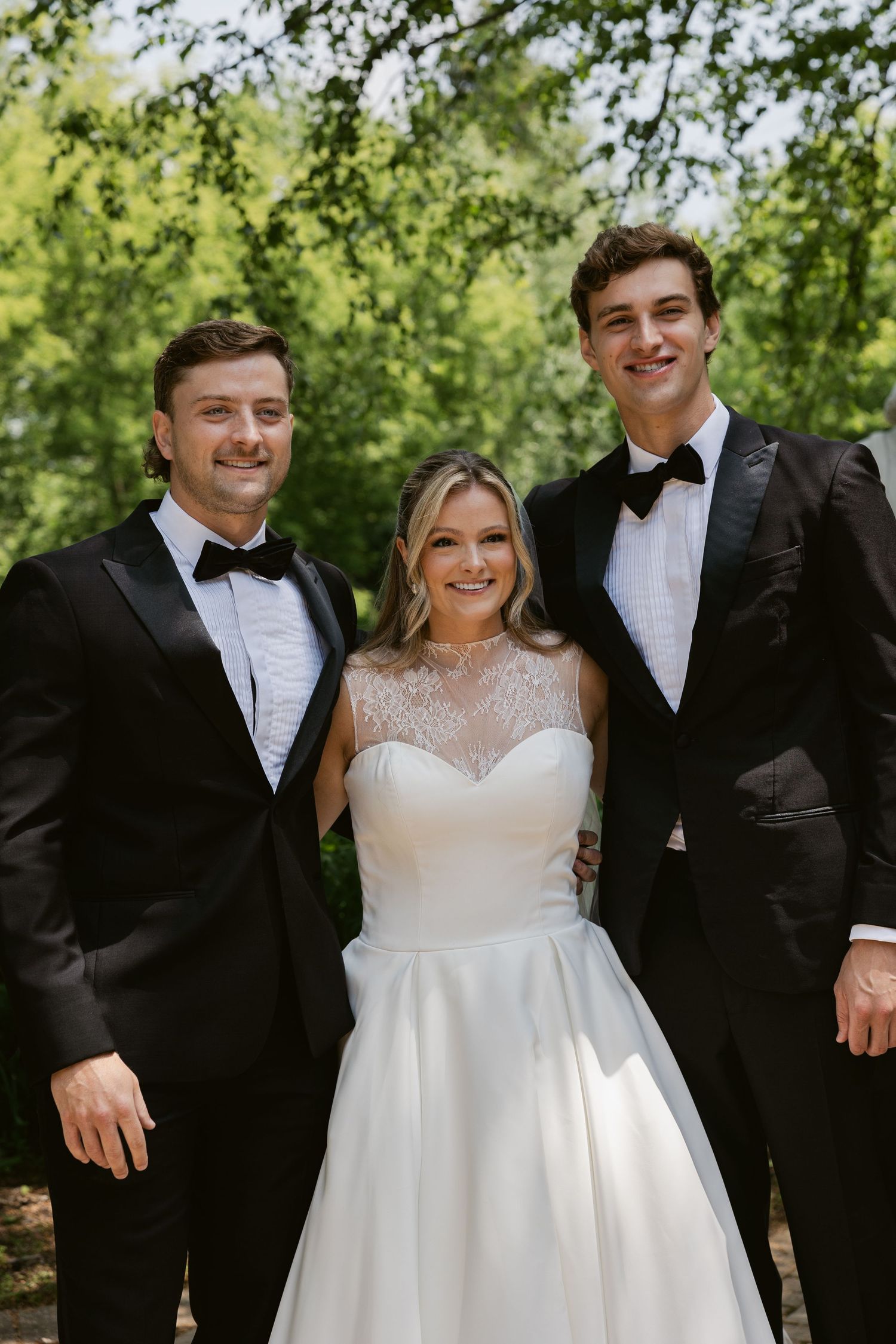 Wedding party members smile together in an outdoor garden setting on a sunny day.
