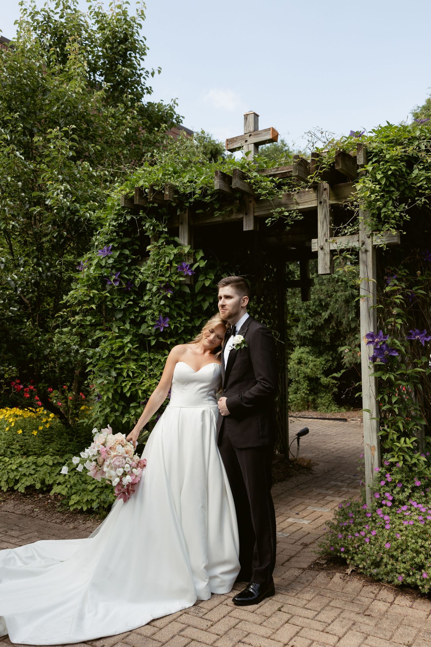Wedding couple standing under wooden garden arbor with climbing vines and flowers.