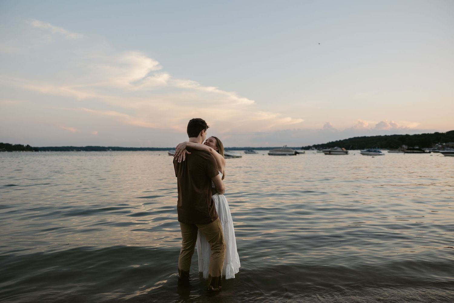 A couple embraces while standing in the water at sunset on a serene lake with hills in the background.