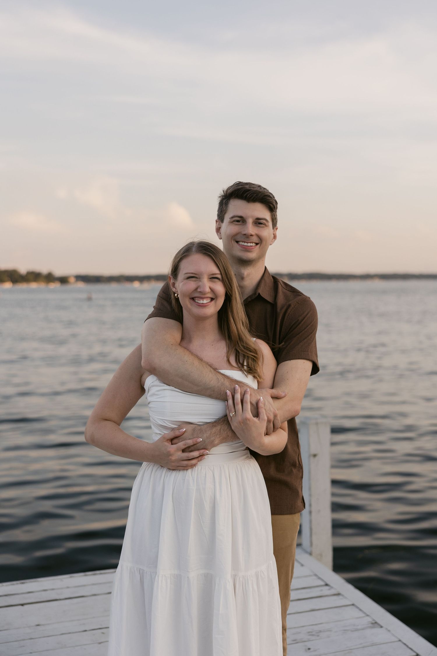 Couple shares intimate embrace on wooden dock with water and sunset in background.