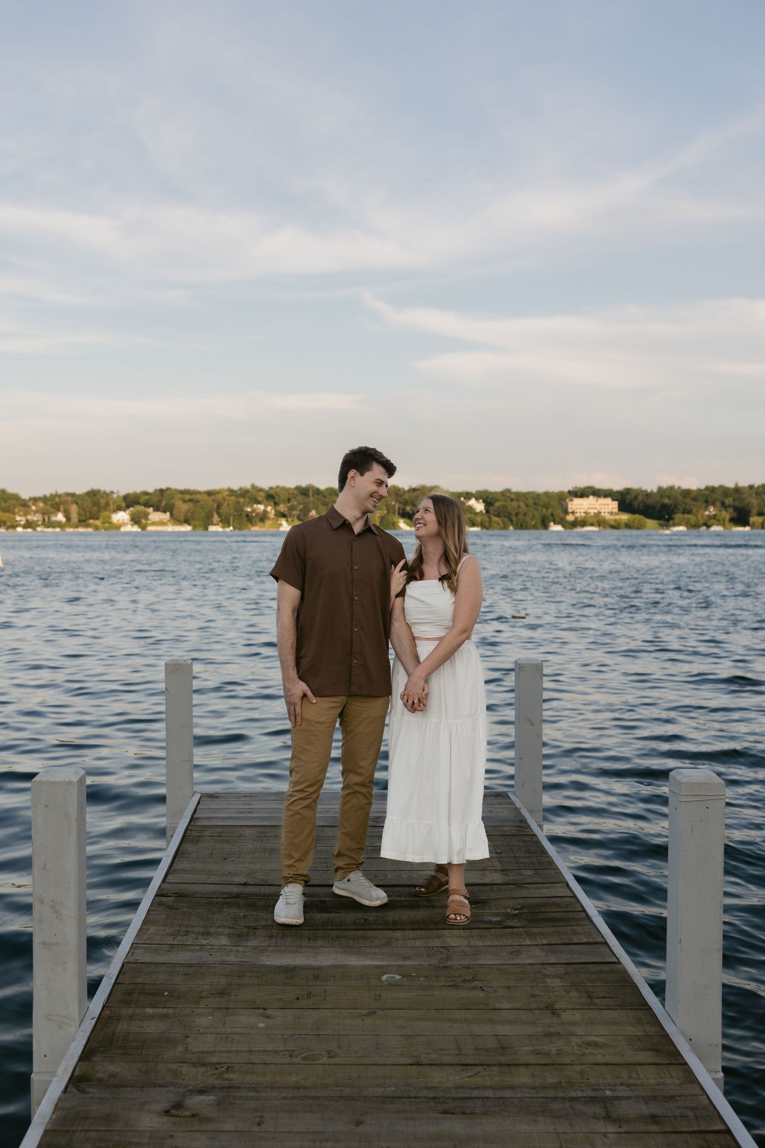 A couple stands together on a wooden dock overlooking a serene lake at sunset.