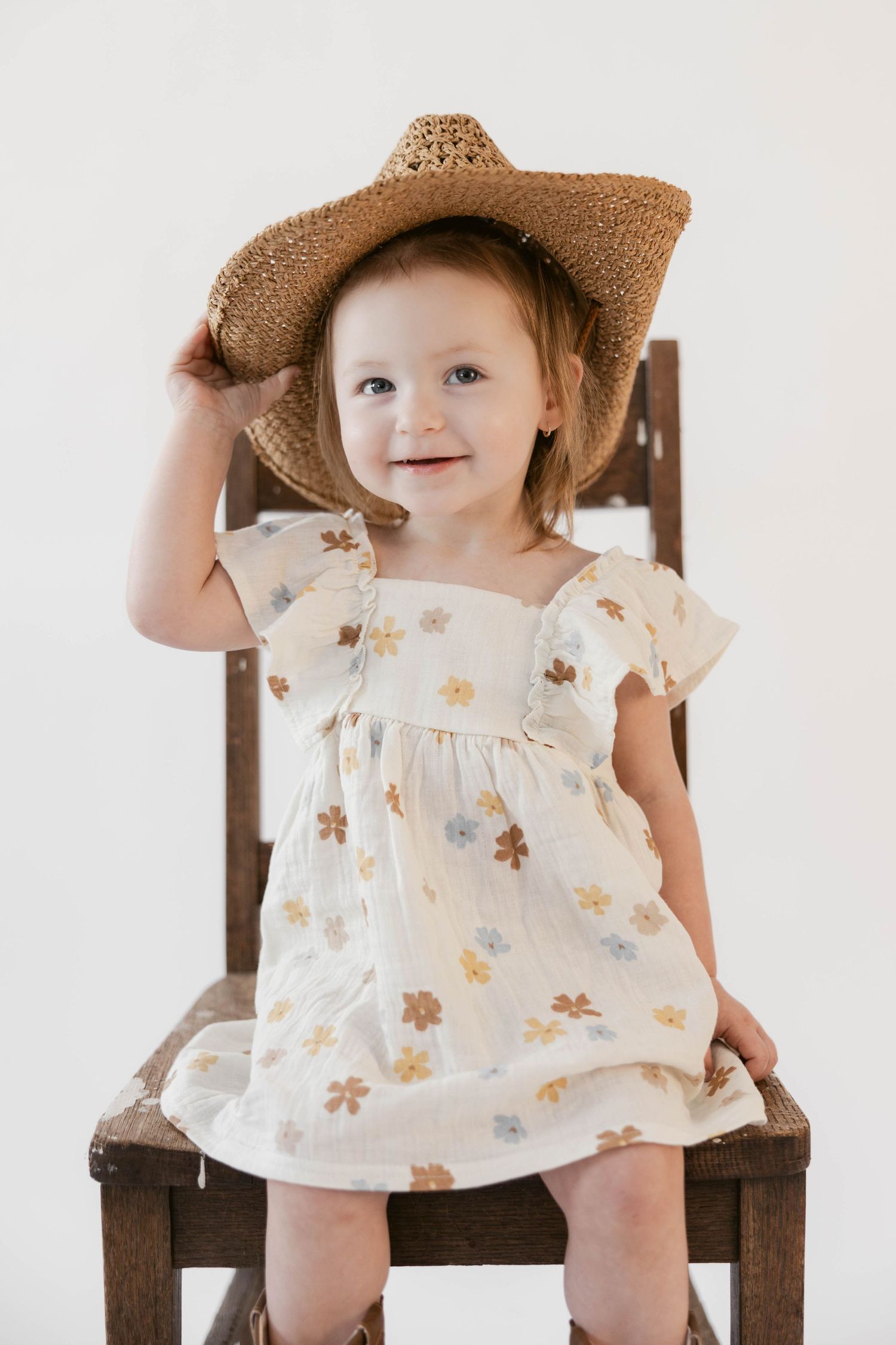 Small child in floral dress and brown straw hat poses cheerfully on wooden chair.