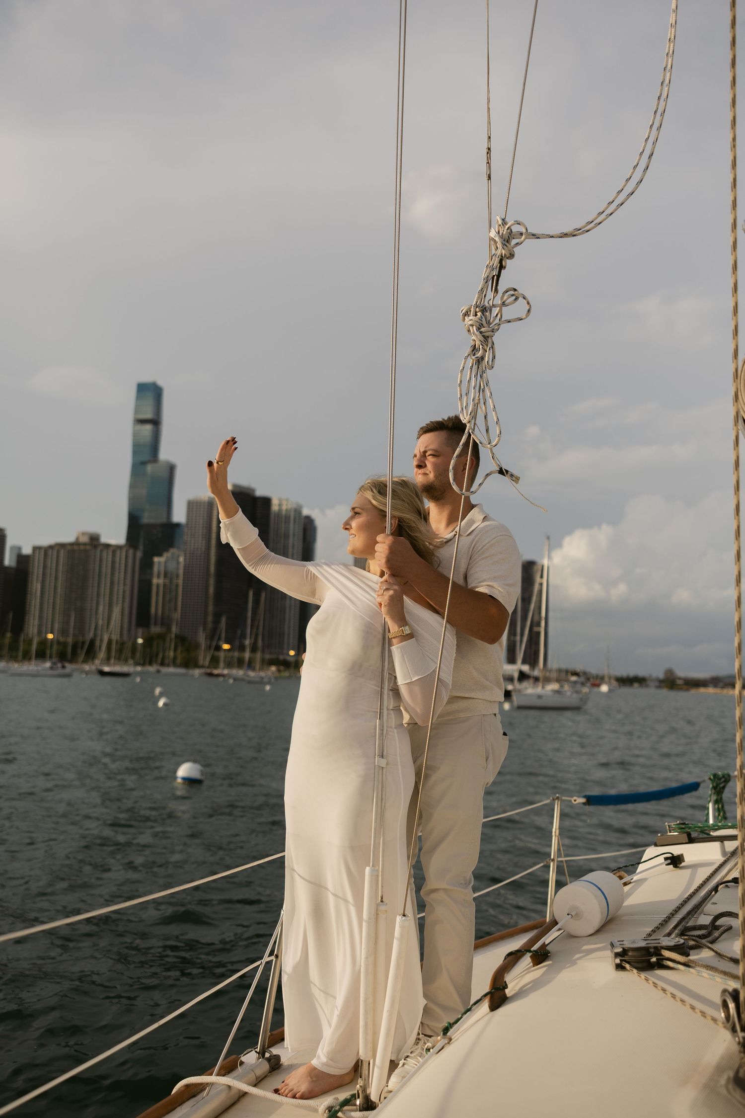 Couple in white outfits celebrating joyfully on sailboat deck with urban waterfront backdrop.