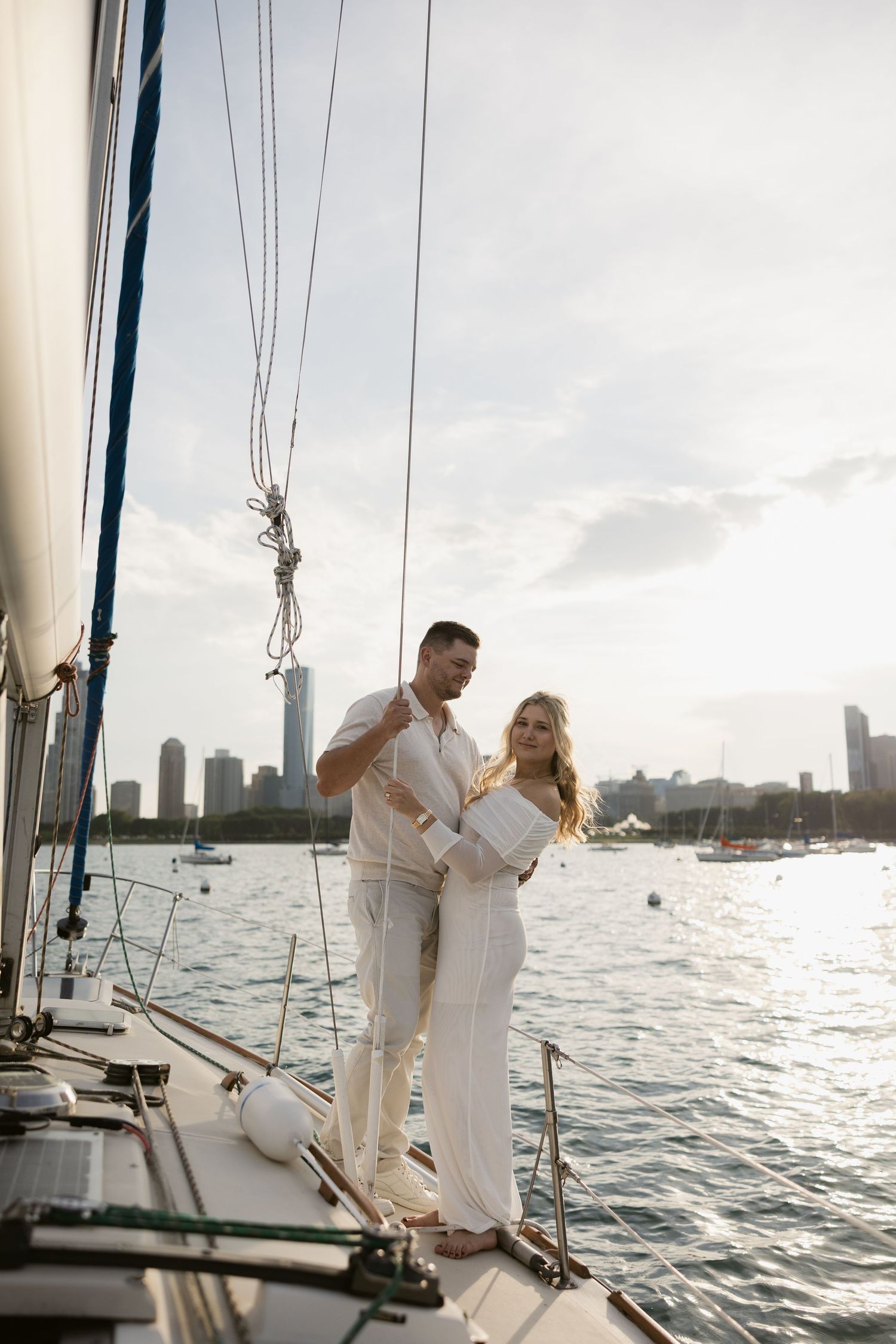 Couple in white outfits sharing intimate moments on a sailboat with city skyline at sunset.