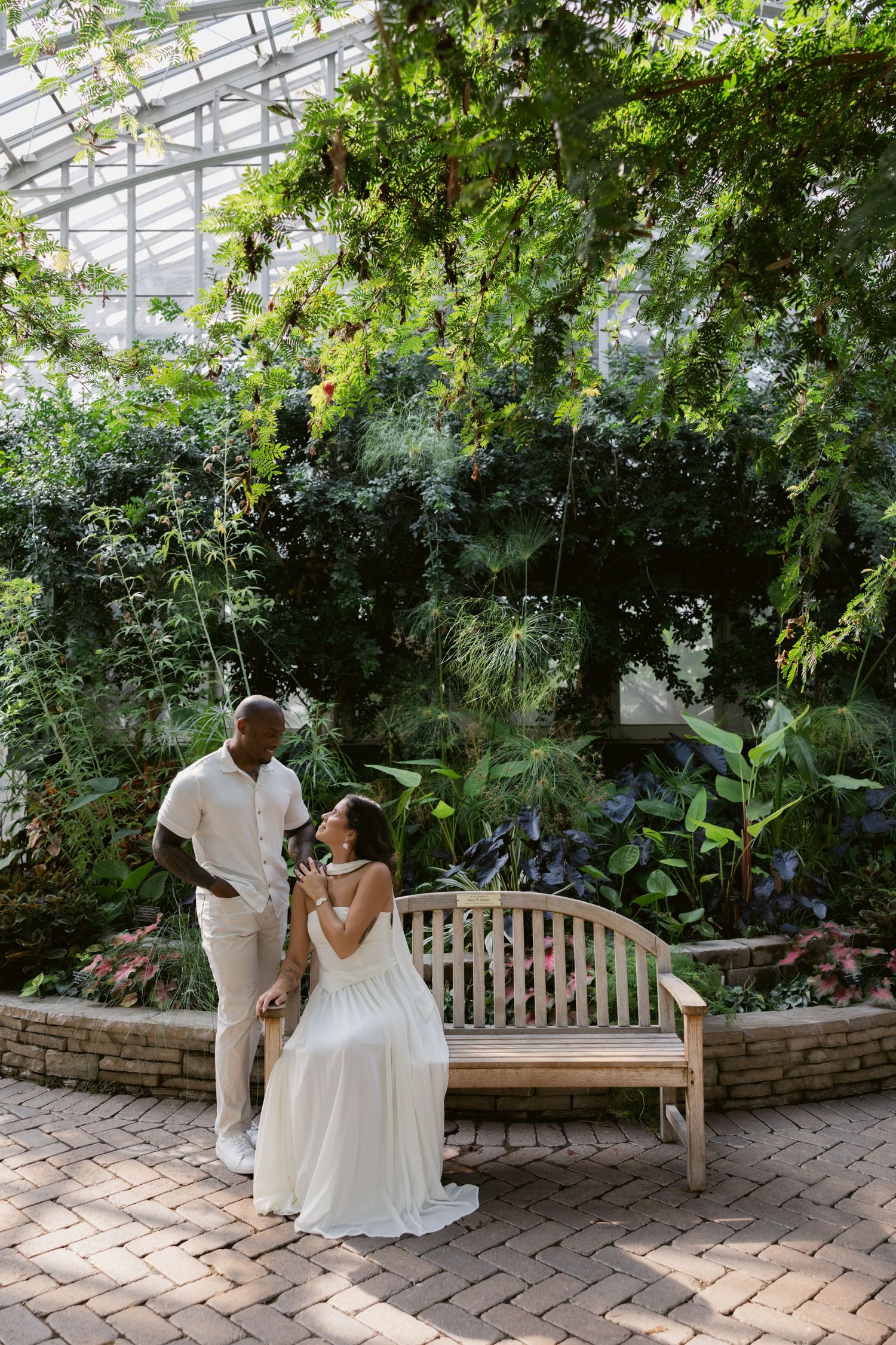 A couple shares a tender moment on a wooden bench in a greenhouse garden setting.
